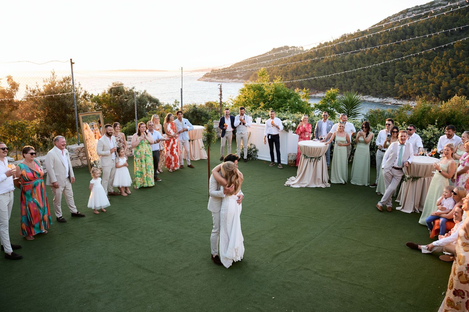 A couple shares their first dance surrounded by wedding guests on an outdoor terrace overlooking coastal hills at sunset.