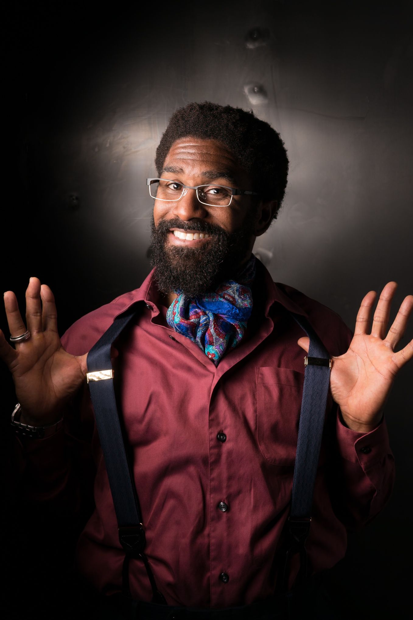 Person in burgundy button-down shirt with navy suspenders and blue bow tie smiling with raised hands against dark background.