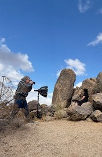 A photographer crouching to take photos of large standing rocks against a blue sky with fluffy white clouds.