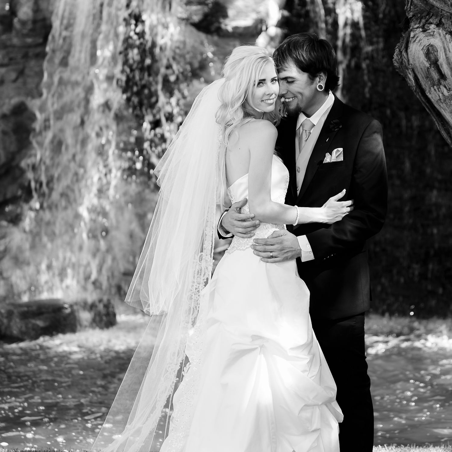 Black and white wedding portrait with couple embracing by cascading waterfall in romantic outdoor setting.