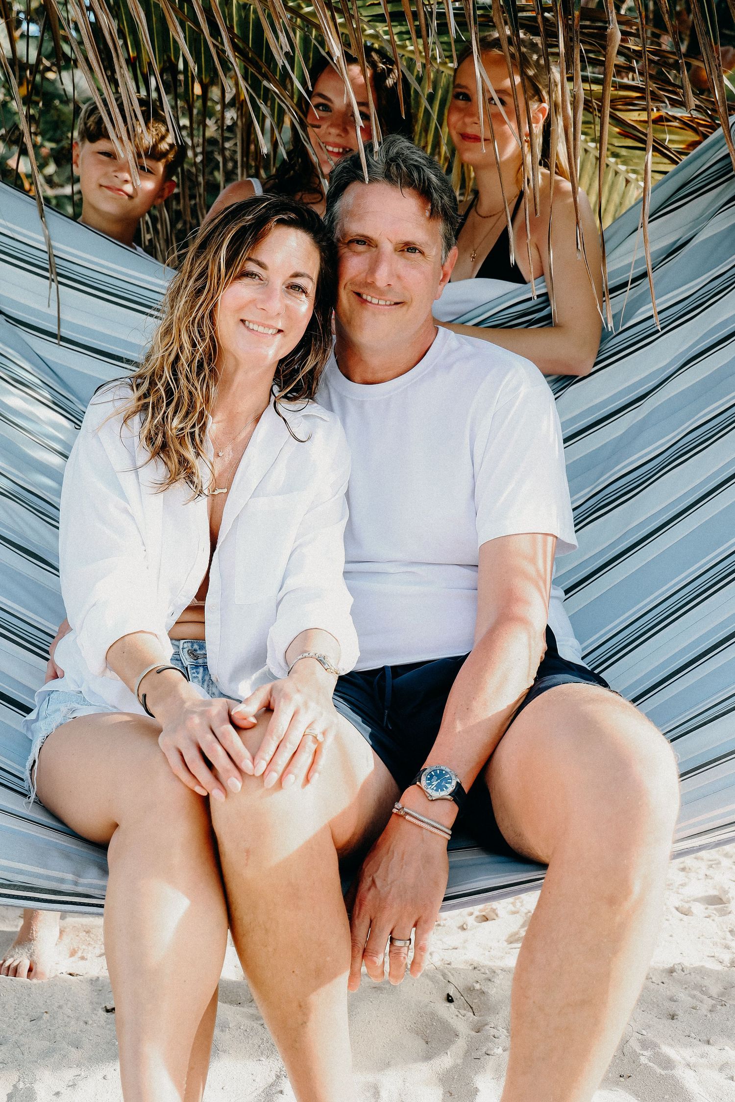 A couple relaxes together in a hammock surrounded by tropical palm fronds on a sunny day.