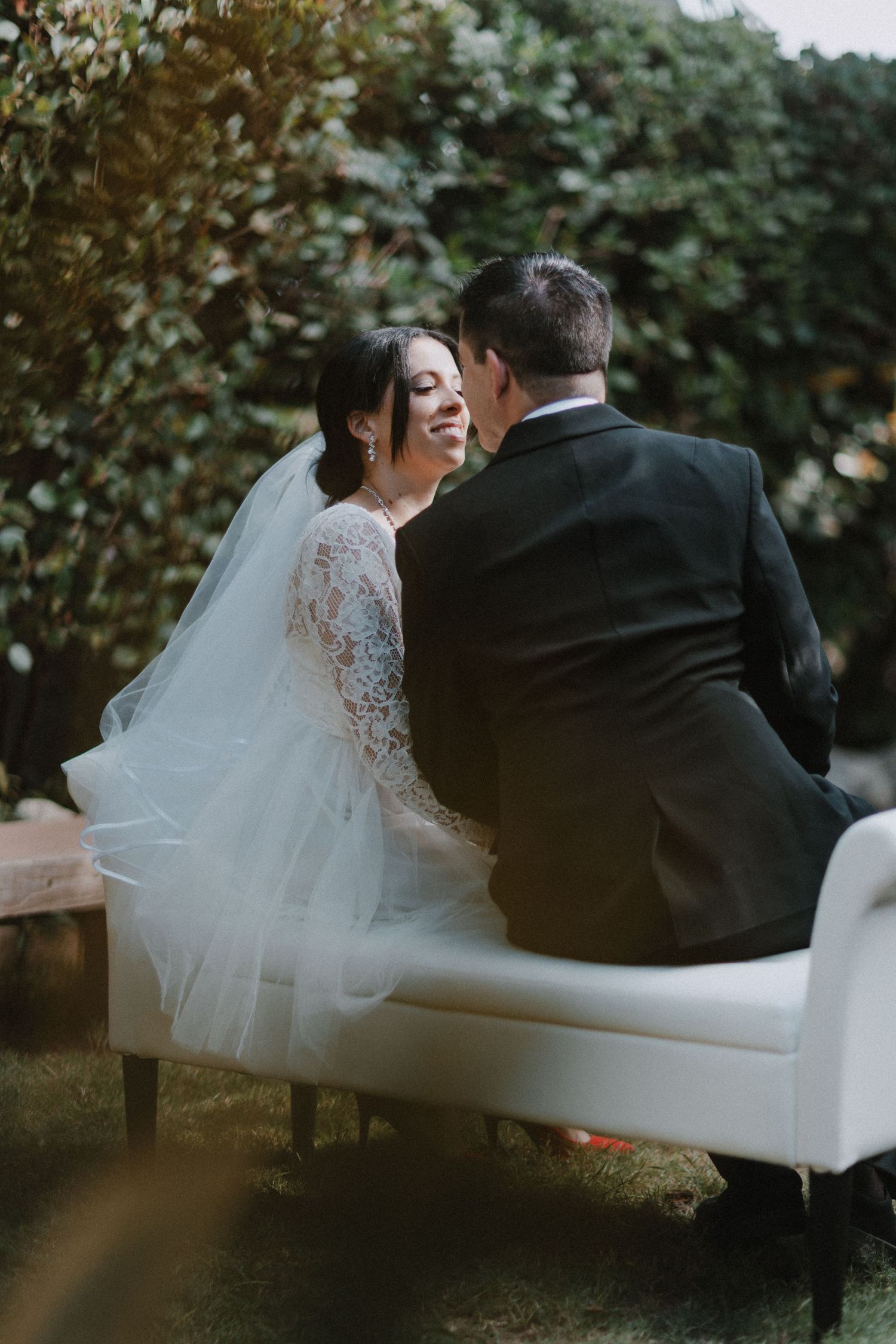 Romantic wedding portrait of couple sitting on white sofa in garden setting.
