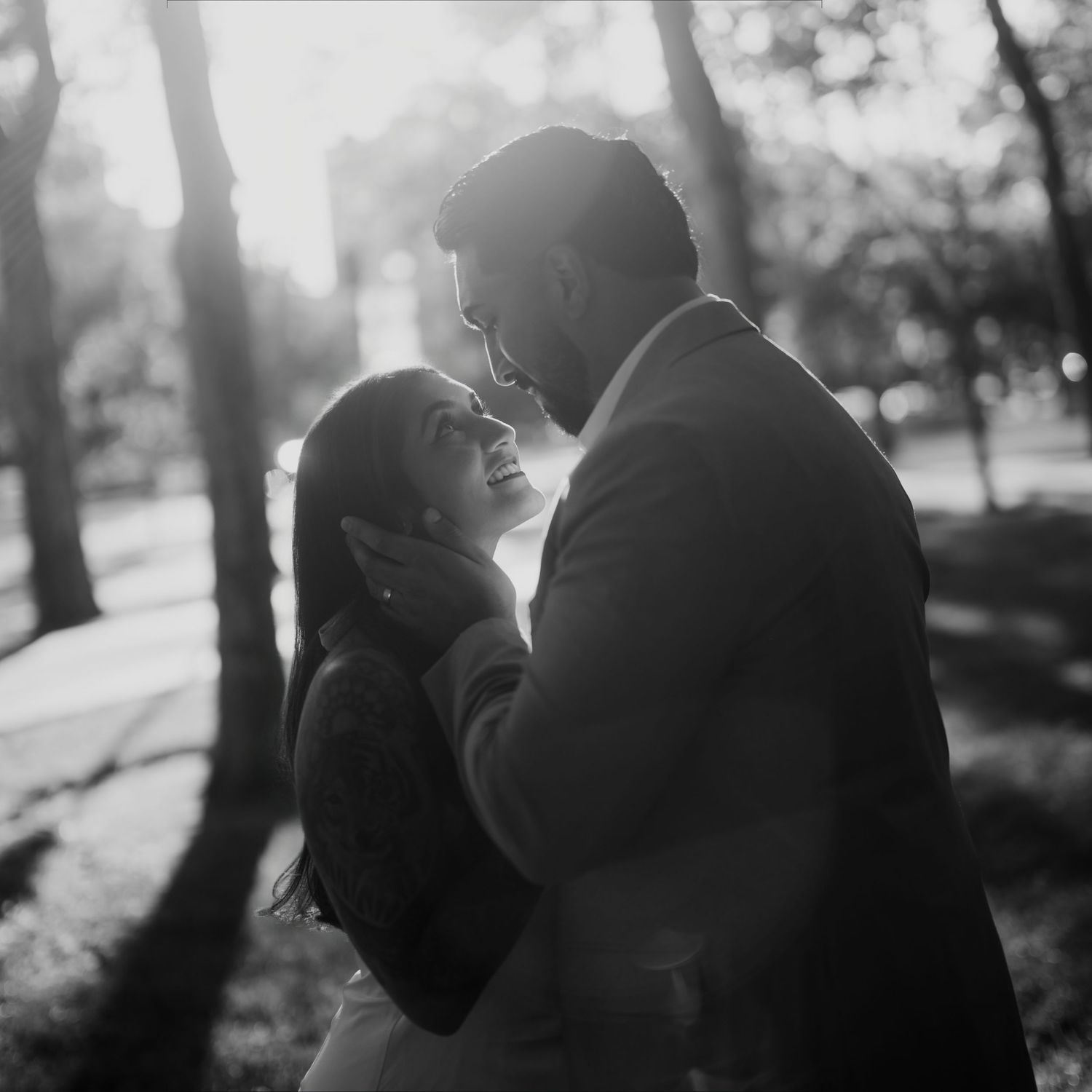 Black and white photo of a couple embracing outdoors, woman smiling up at man in soft backlit park setting.