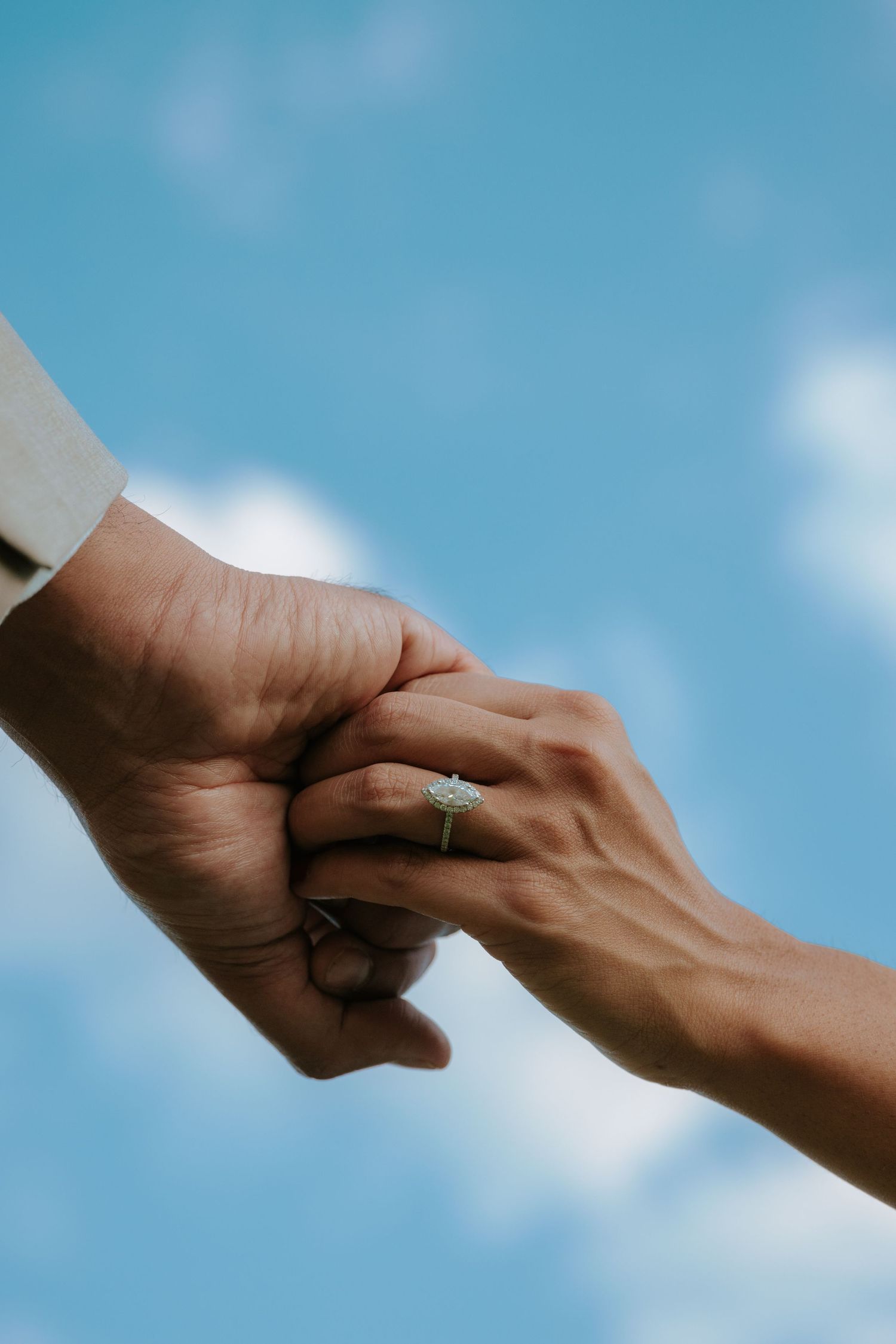 Two hands holding together against a blue sky, with a diamond engagement ring on the woman's finger.