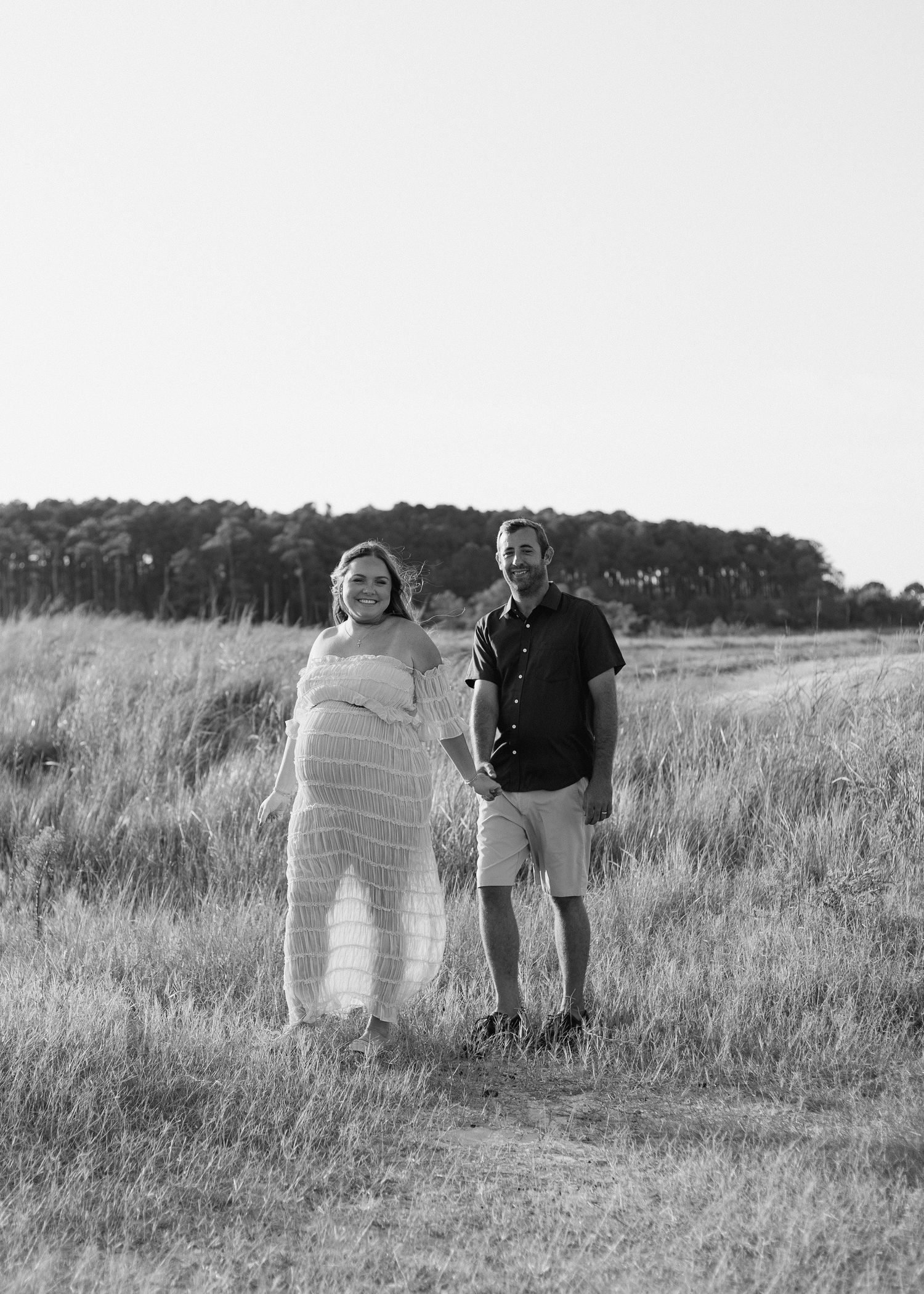 Black and white photo of a pregnant couple holding hands in a grassy field with trees in the background.