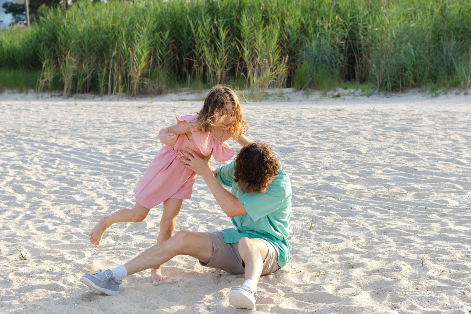 Two people share a playful moment together on a sandy beach with tall grass in the background.