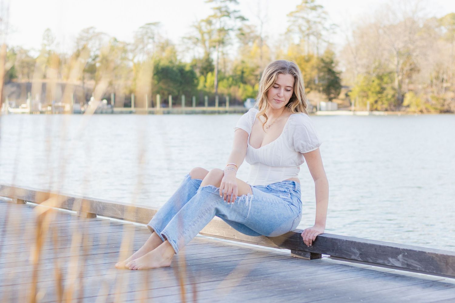 Someone in casual attire poses on a wooden dock by a serene lake on a sunny day.
