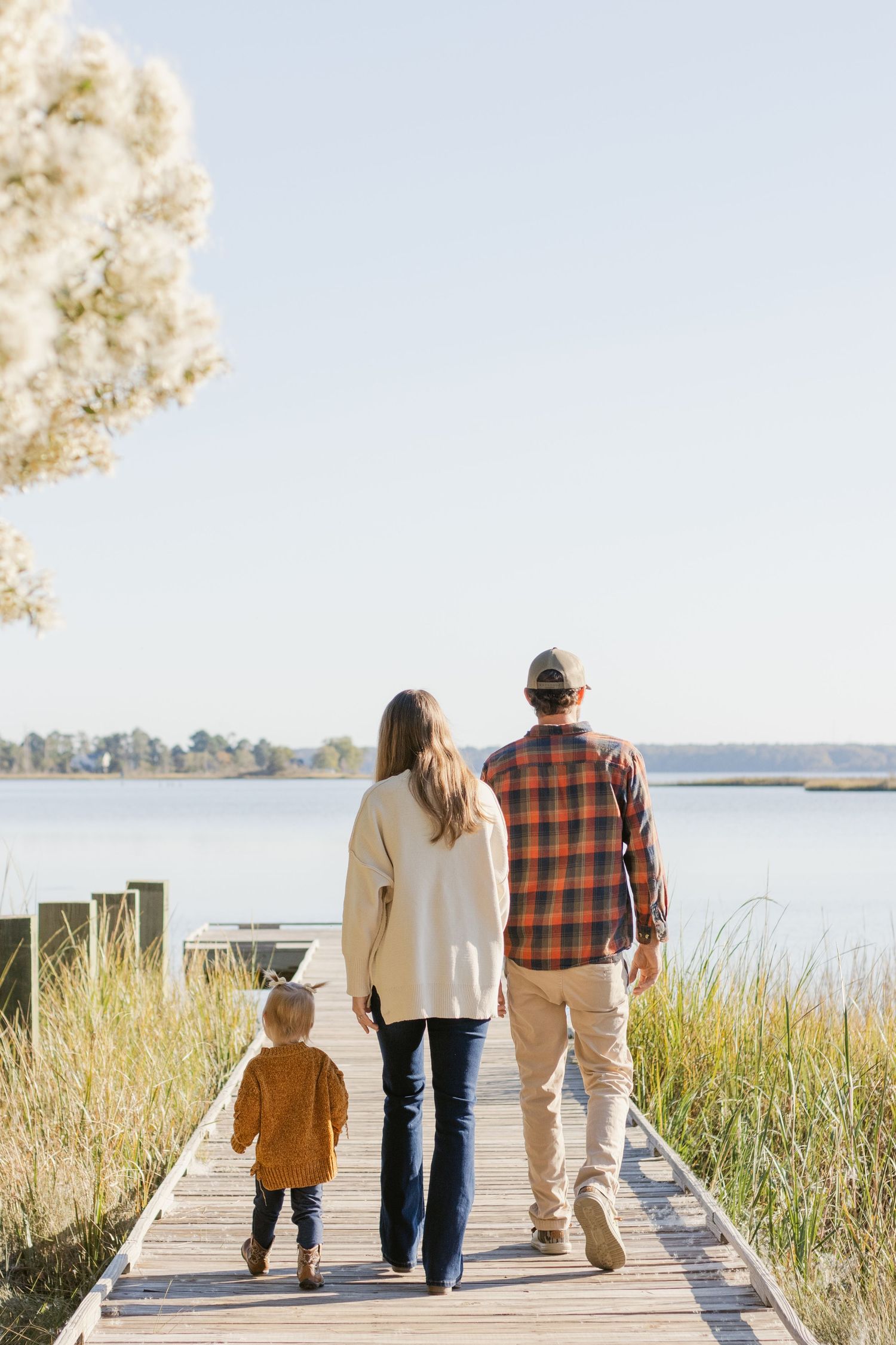 A family walks together on a wooden boardwalk through marsh grass beside a serene lake on a sunny autumn day.