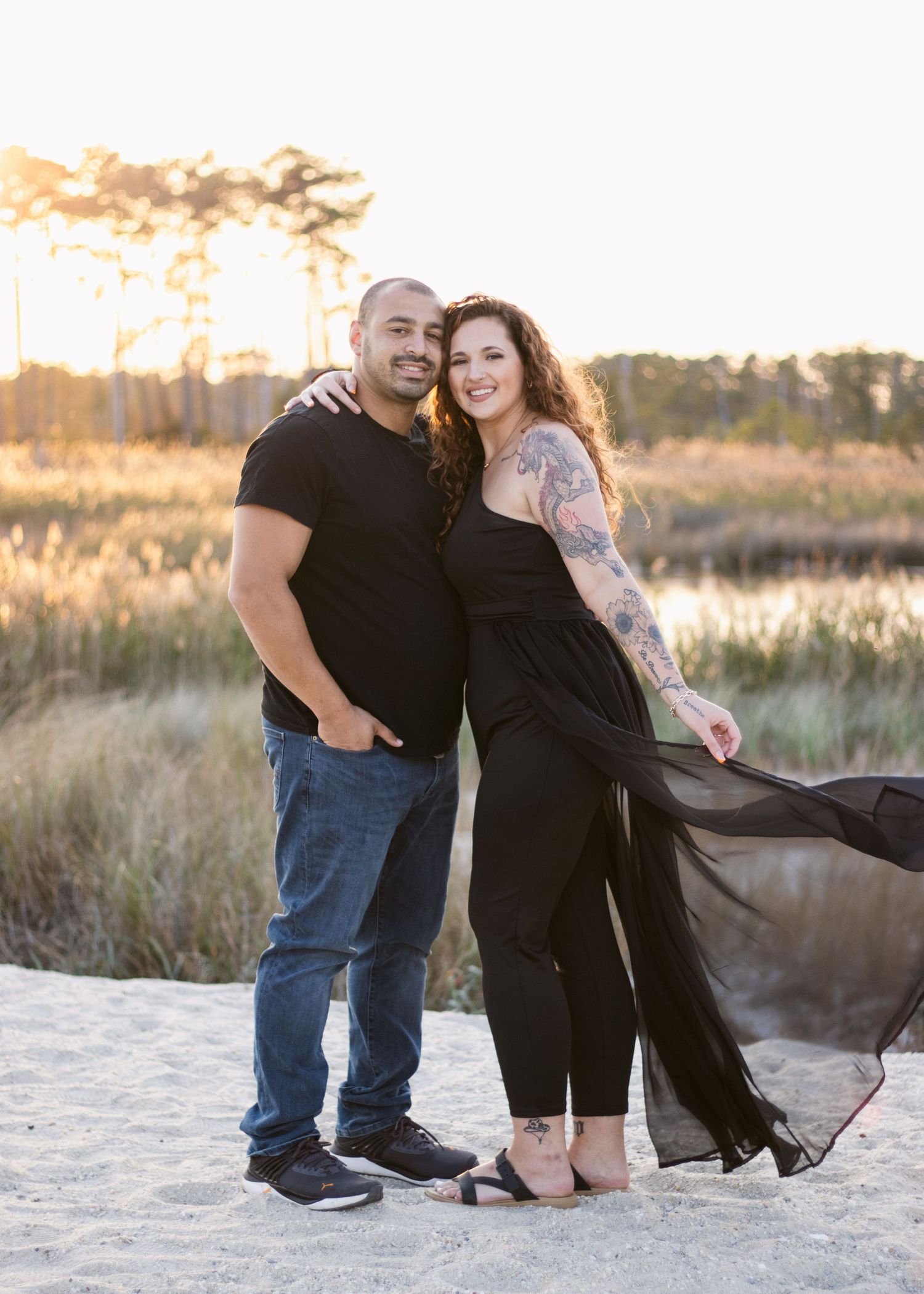 A couple stands together at sunset on the beach with flowing black dress and casual attire.