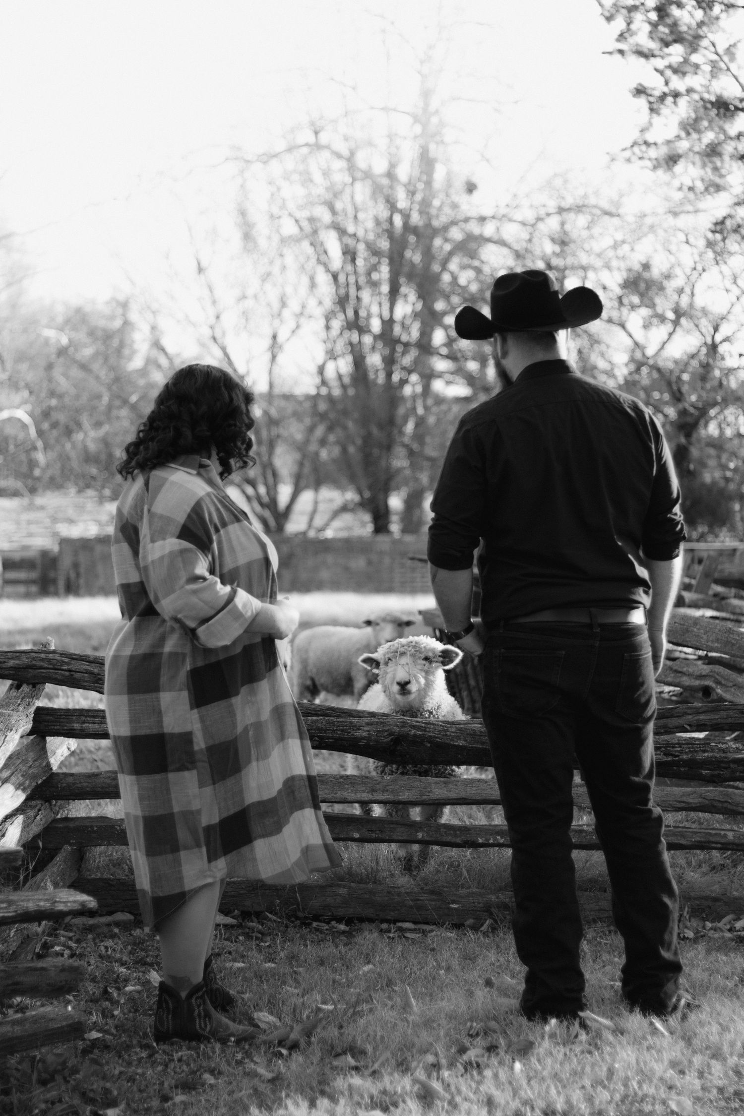 A couple in rustic attire stands by a wooden fence in monochrome, with one wearing a cowboy hat.