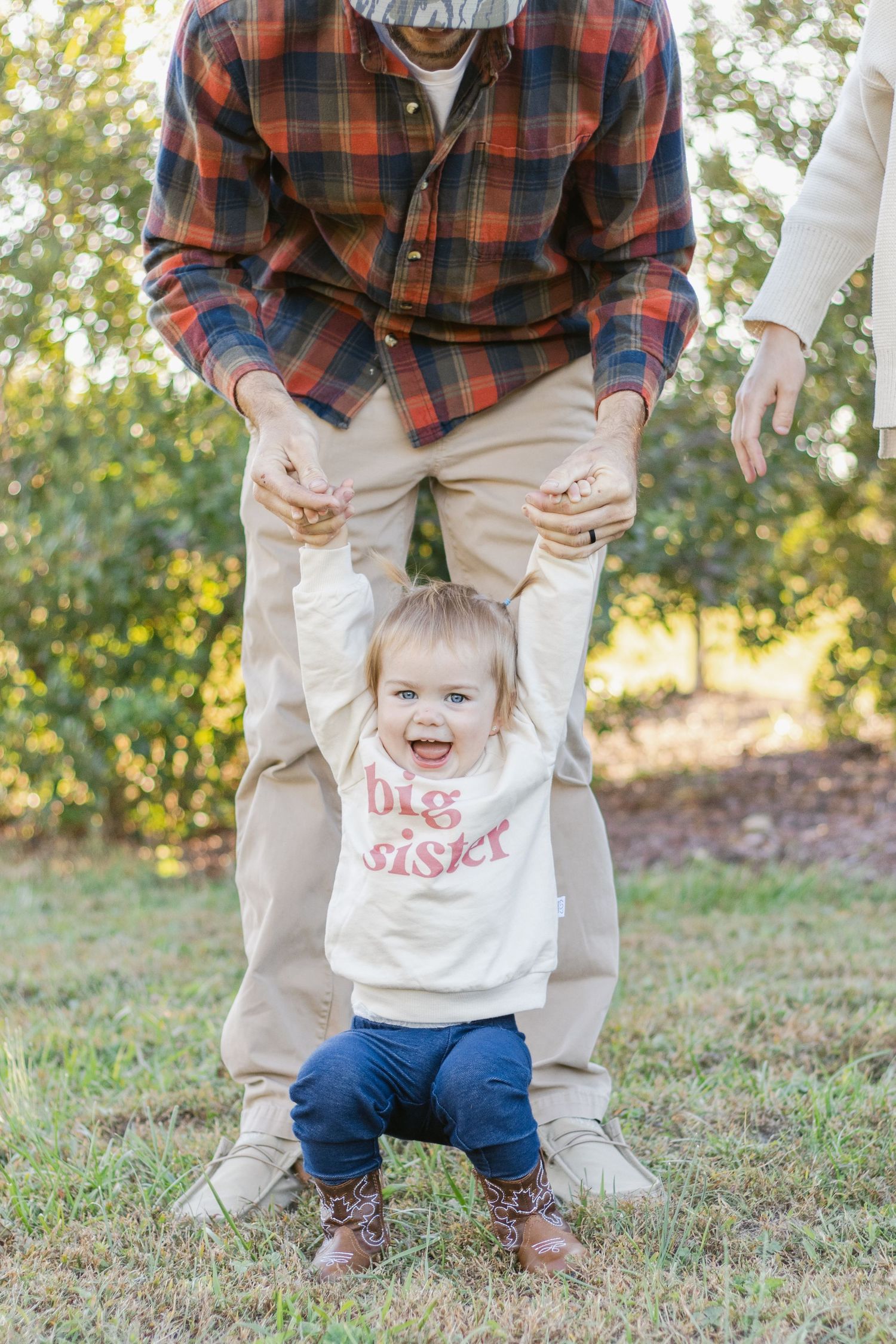Toddler in Big Sister sweater learning to walk with support outdoors.