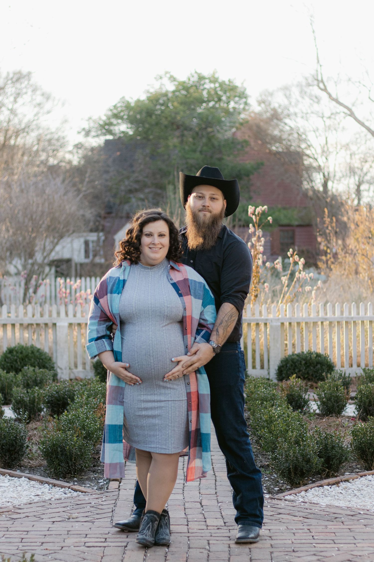 A couple stands together on a brick pathway with a white picket fence and garden in the background.