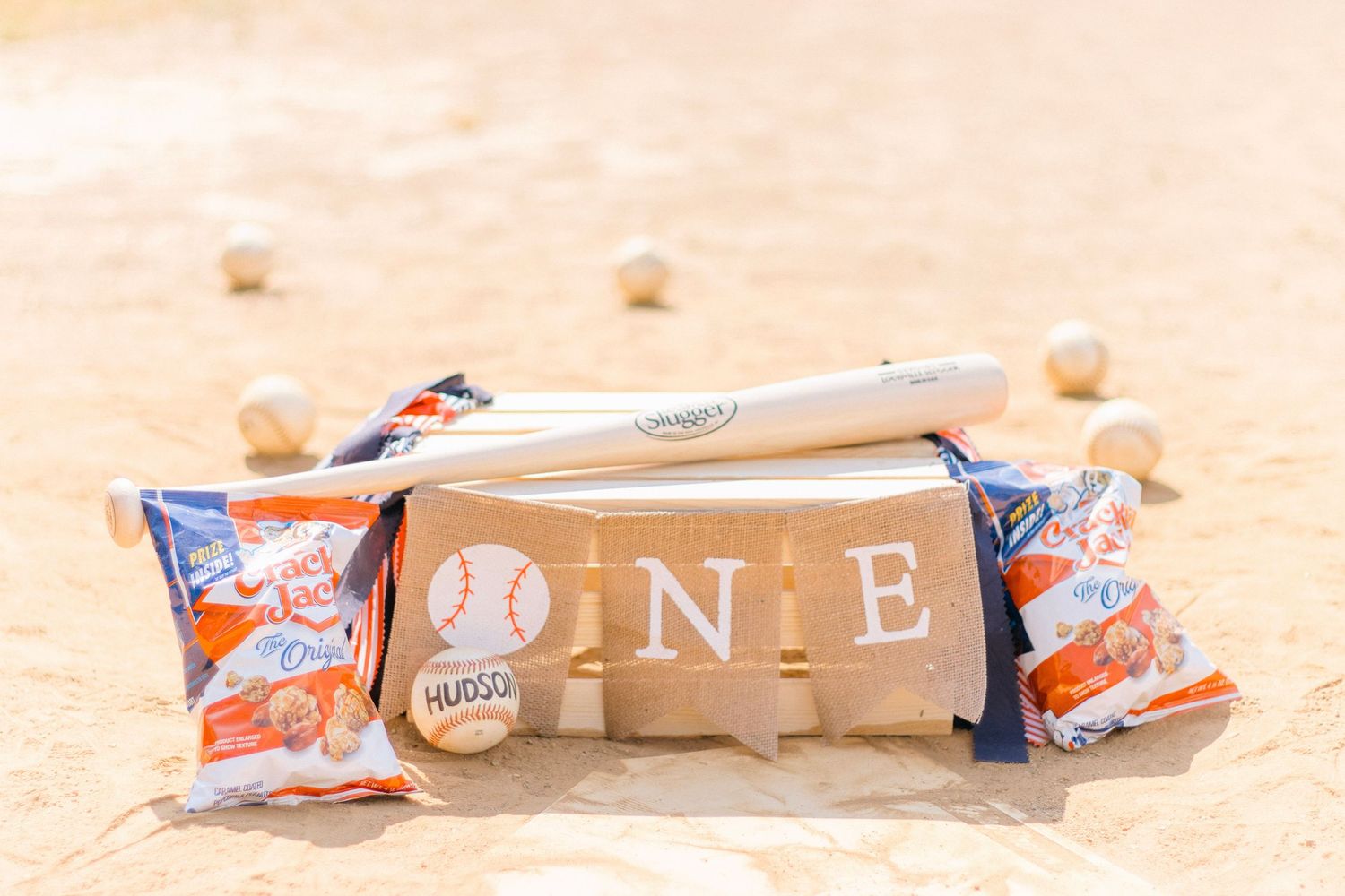 A baseball themed first birthday party decoration with wooden blocks spelling 'ONE' and Cracker Jack boxes on sand.