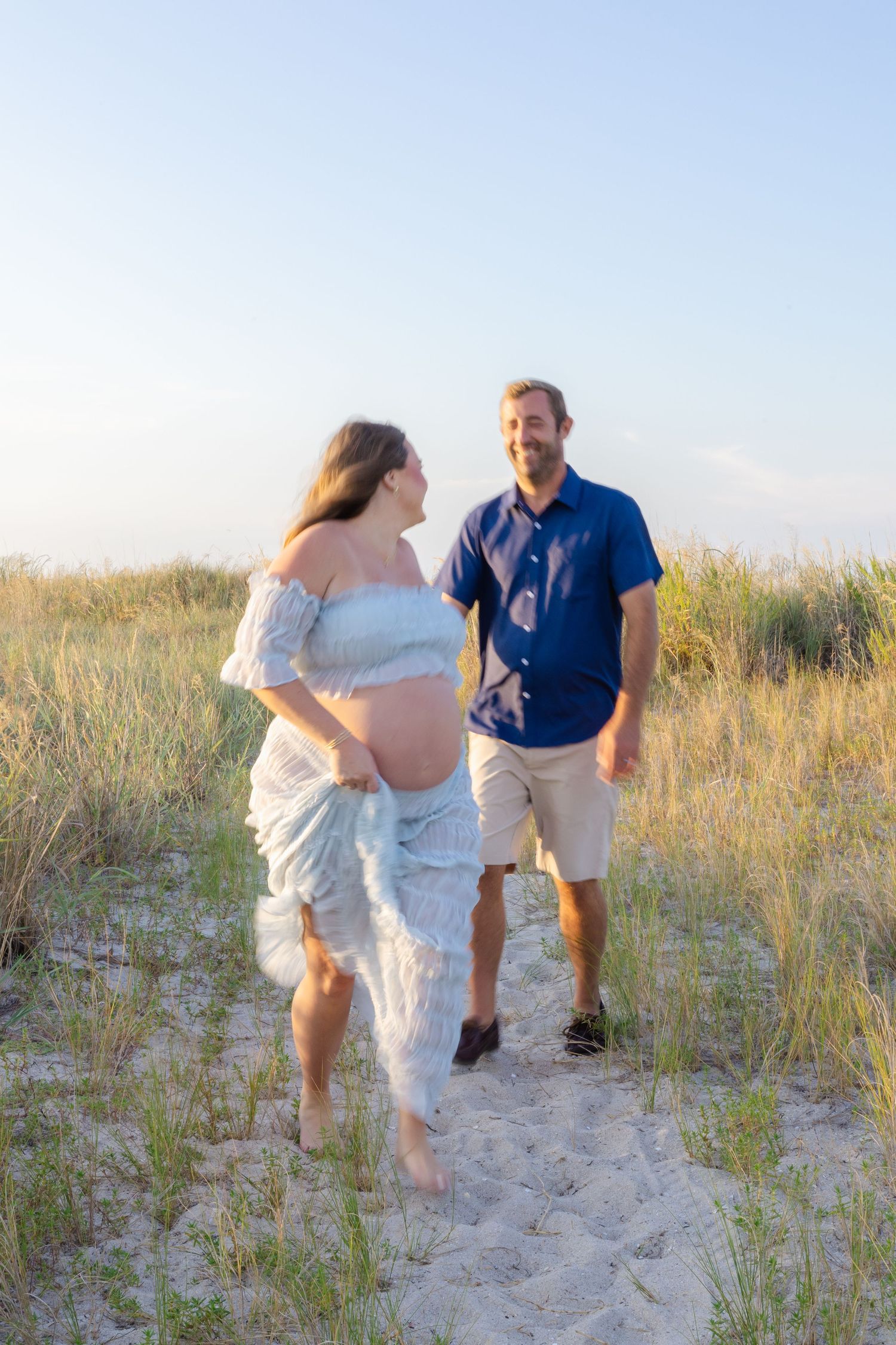Pregnant woman in white lace dress walks with man in blue shirt on sandy beach path surrounded by tall grass.