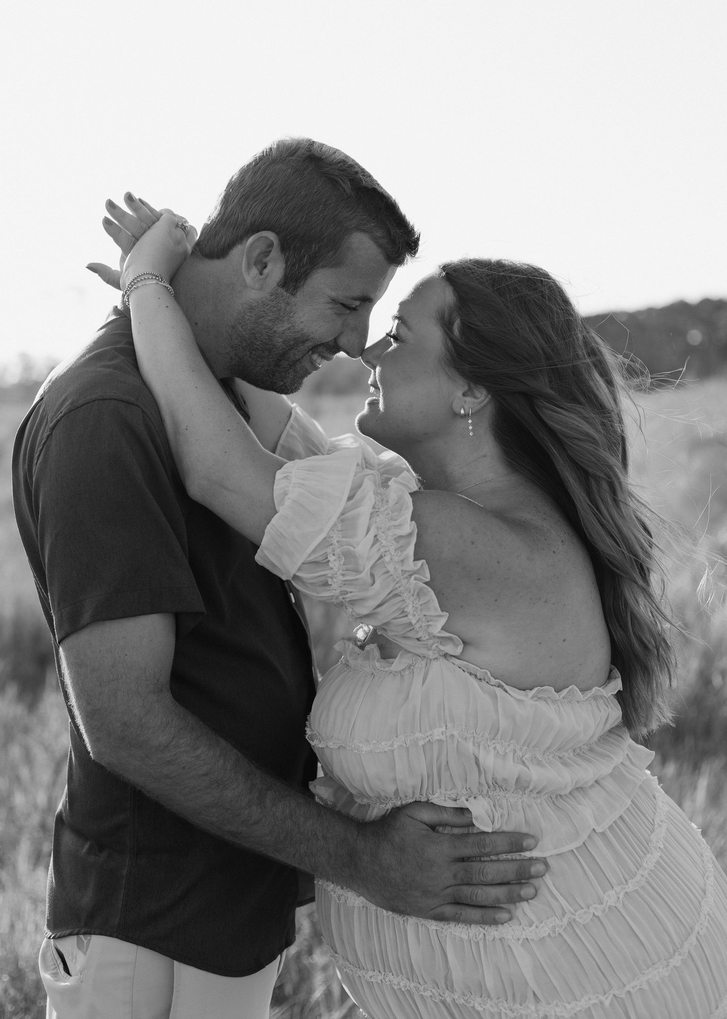 Black and white photo of a couple embracing outdoors, woman in ruffled dress, man in dark shirt, sharing a romantic moment.