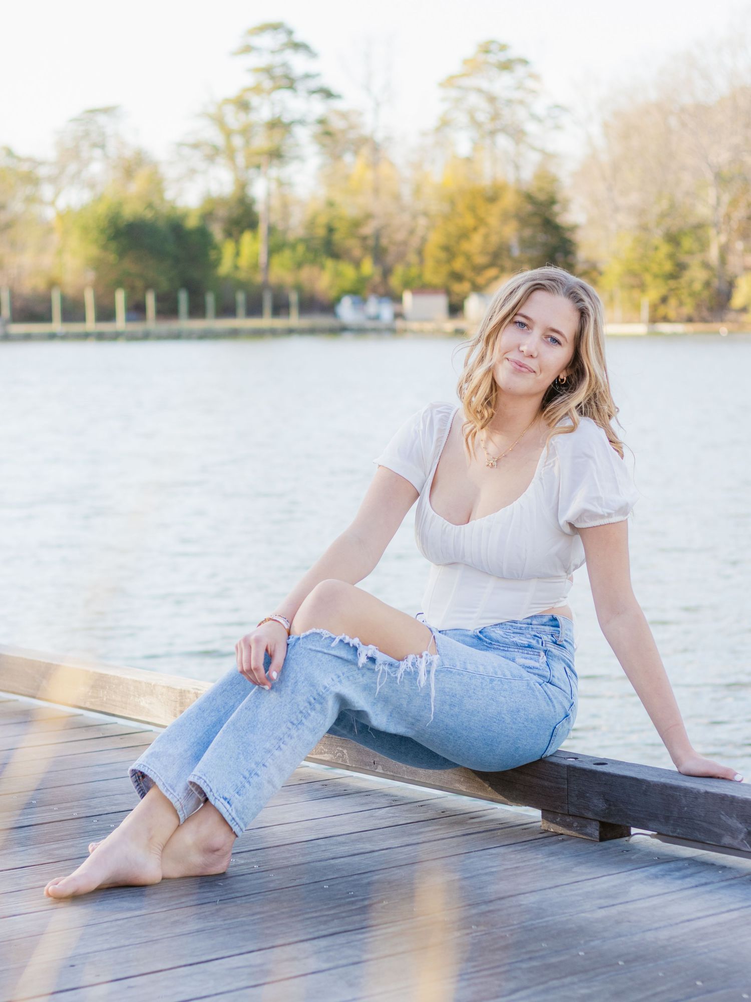 Subject in white top and jeans poses casually on a wooden dock by a scenic lake during golden hour.