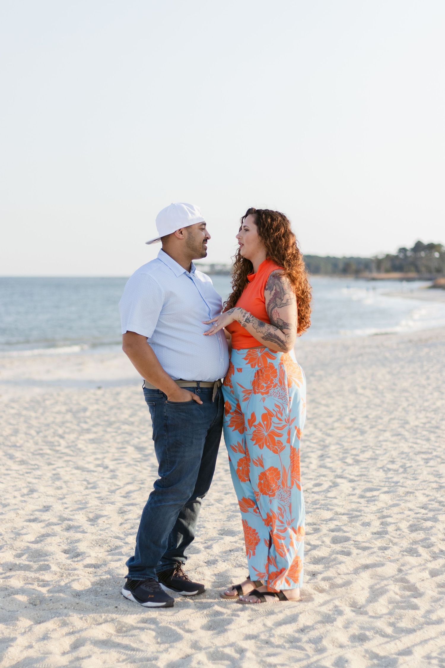A couple shares a tender moment while standing on a sandy beach at sunset, with ocean waves in the background.