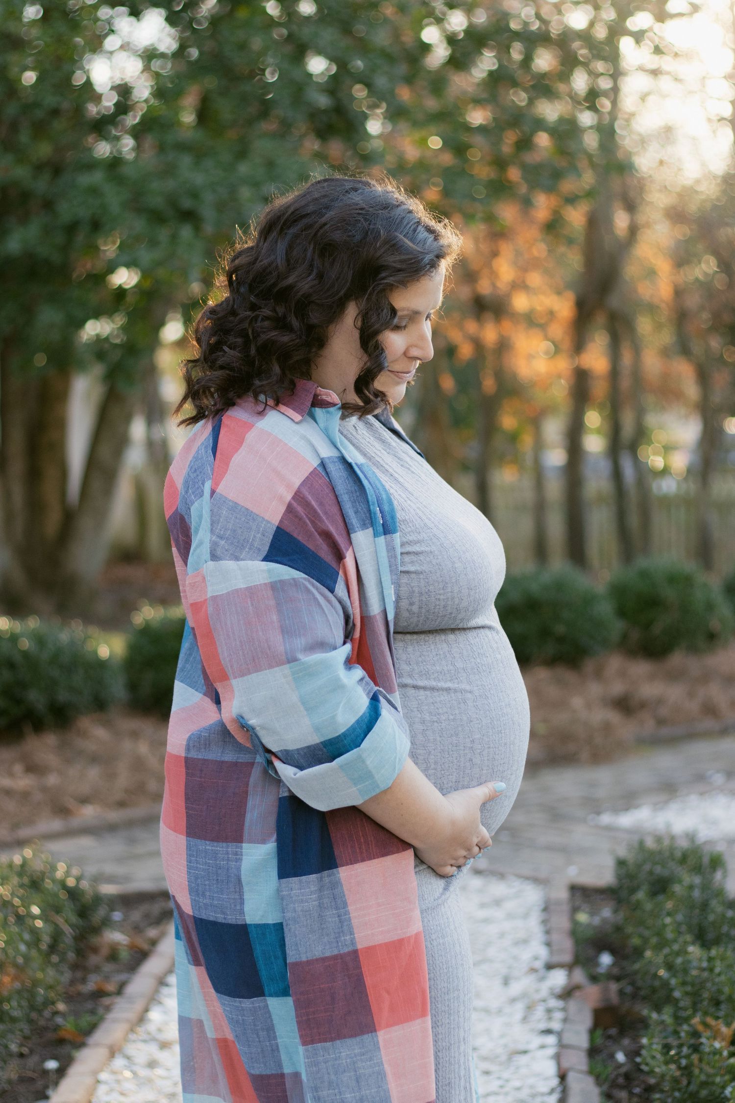 A person in a plaid shirt gently cradles their baby bump while standing on a garden path during sunset.