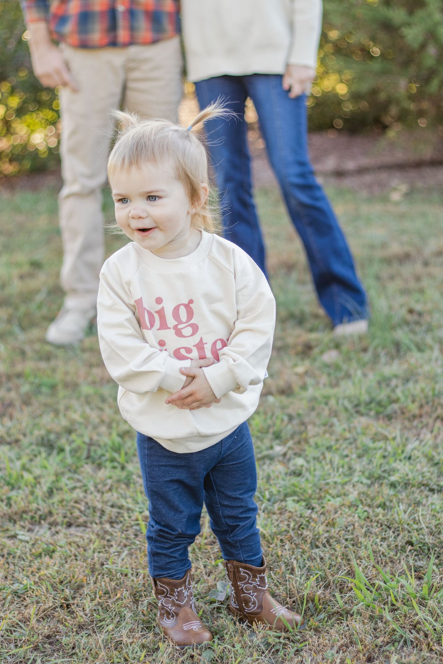 A toddler wearing a white sweater and jeans plays on grassy ground while adults stand in the background.