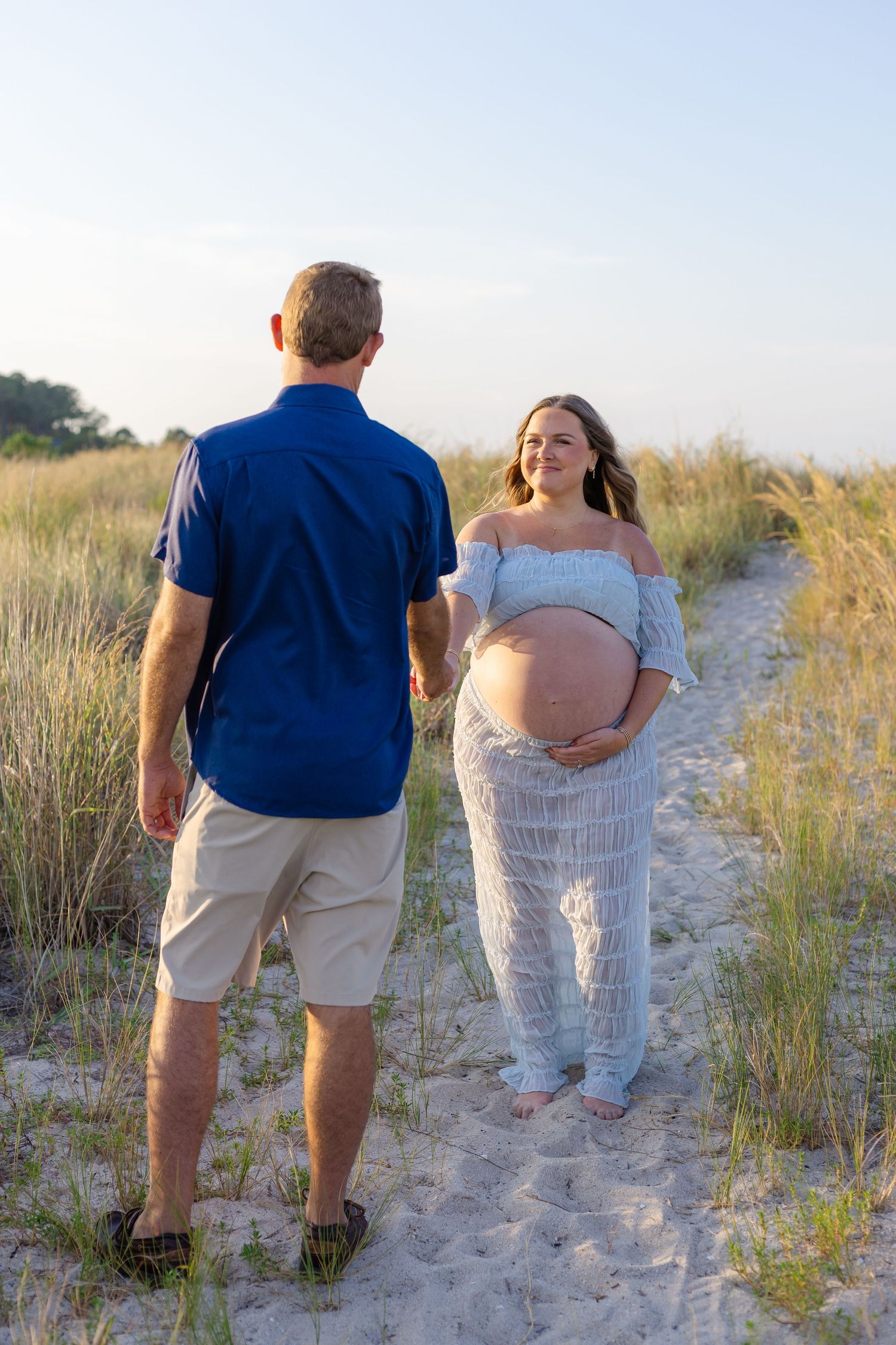 Pregnant woman in white lace outfit holds her baby bump while holding hands with a man on a sandy beach path.
