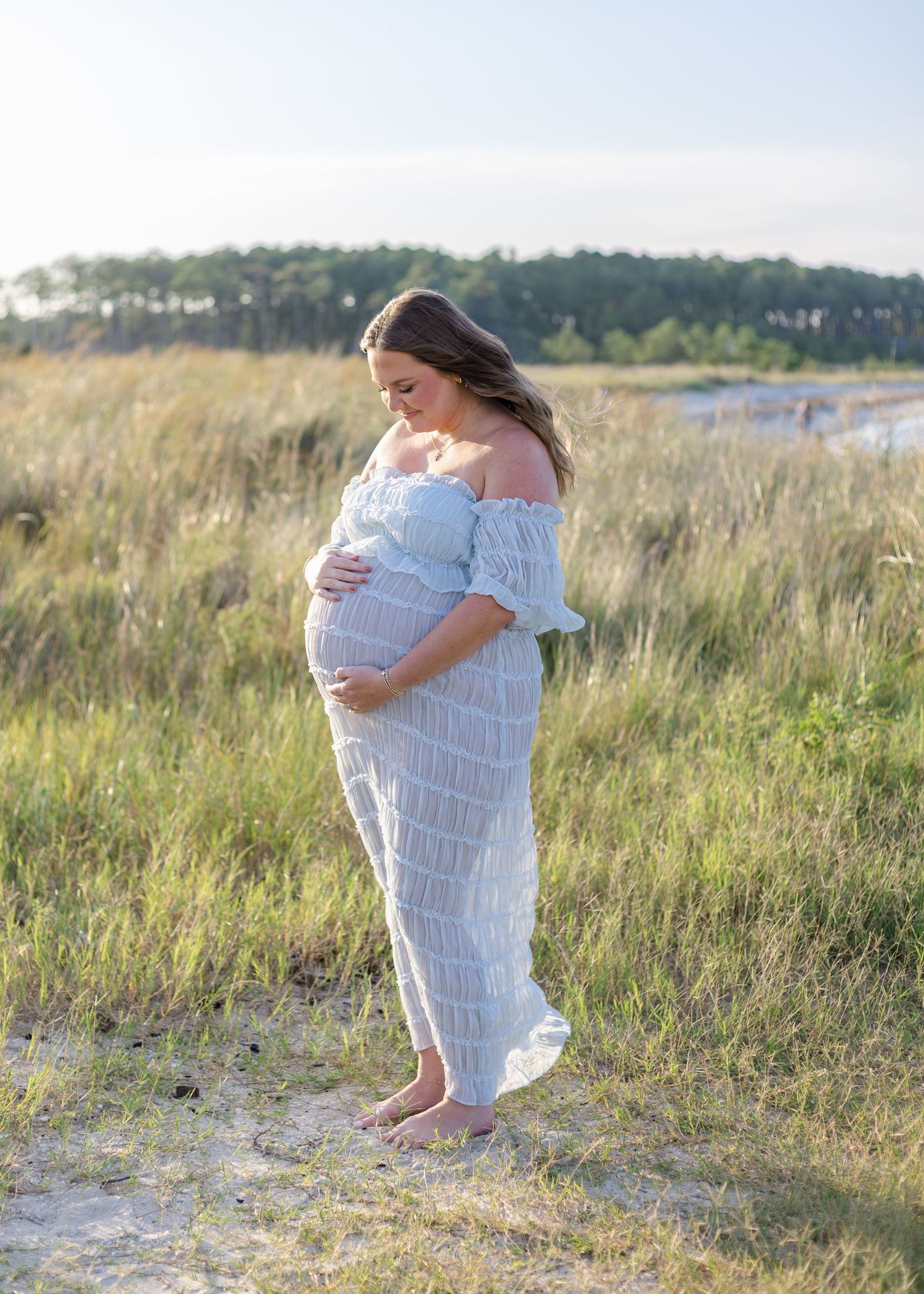 Pregnant woman in white off-shoulder dress cradling baby bump in a sunlit coastal meadow.