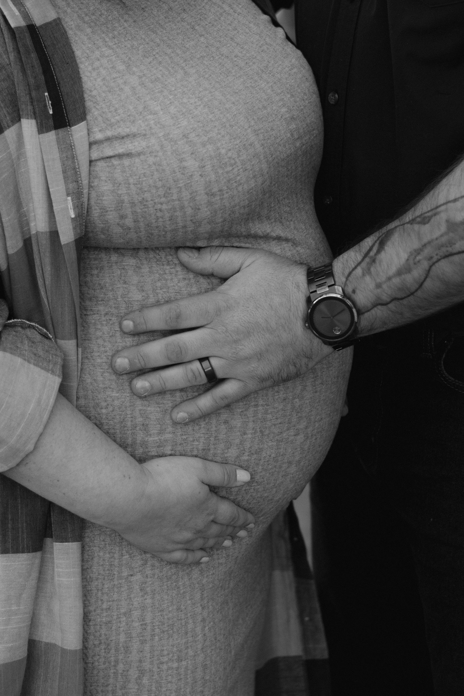 Black and white maternity photo showing hands cradling a pregnant belly, with a wedding ring and watch visible.