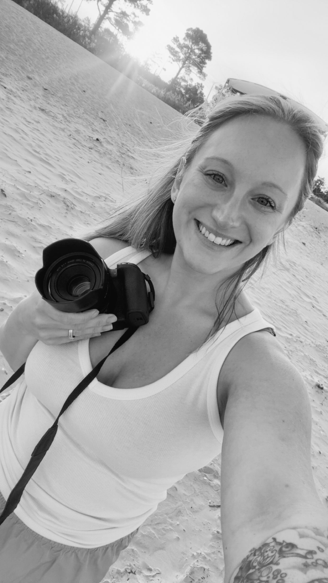 Black and white selfie of someone holding a camera at the beach on a sunny day while wearing a white tank top.