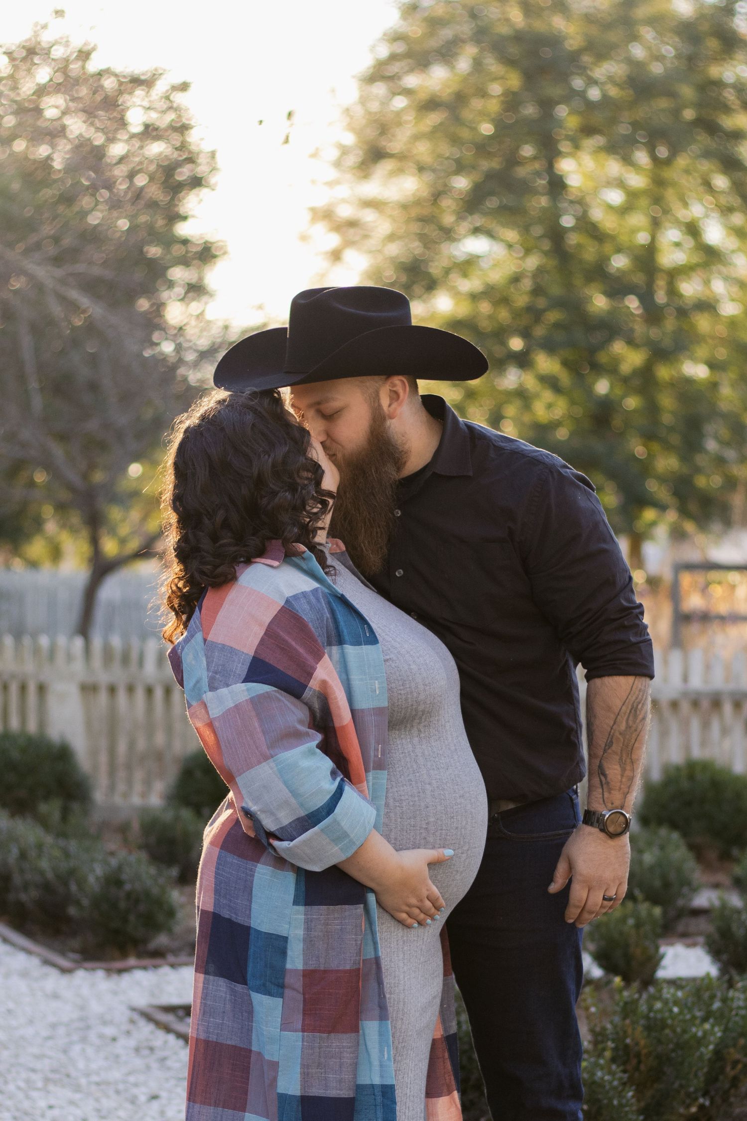 A couple shares a tender moment in a garden at sunset, with a white picket fence and trees in the background.