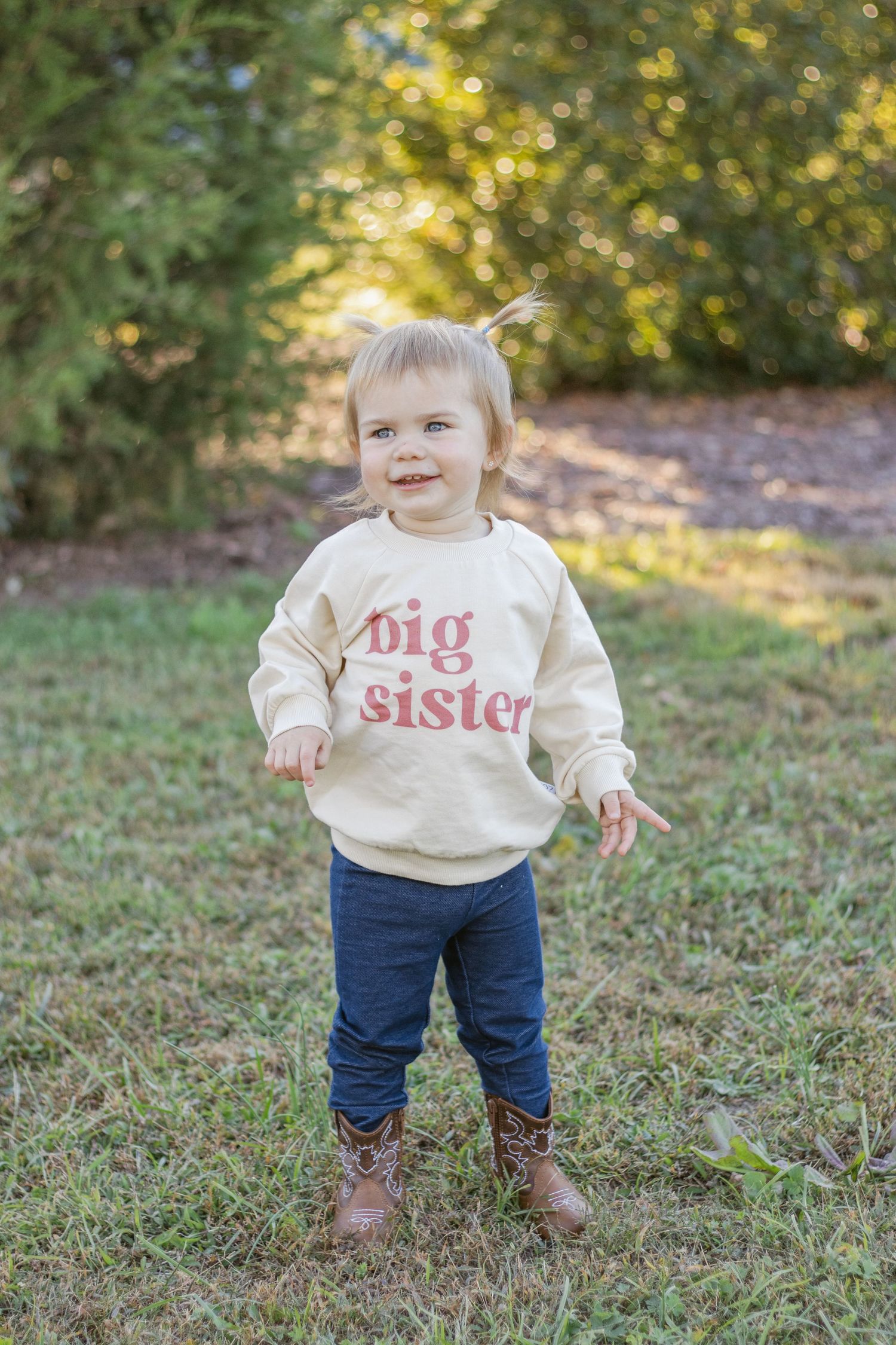 Small child standing outdoors wearing a cream colored 'big sister' sweatshirt, jeans and brown boots on grass.