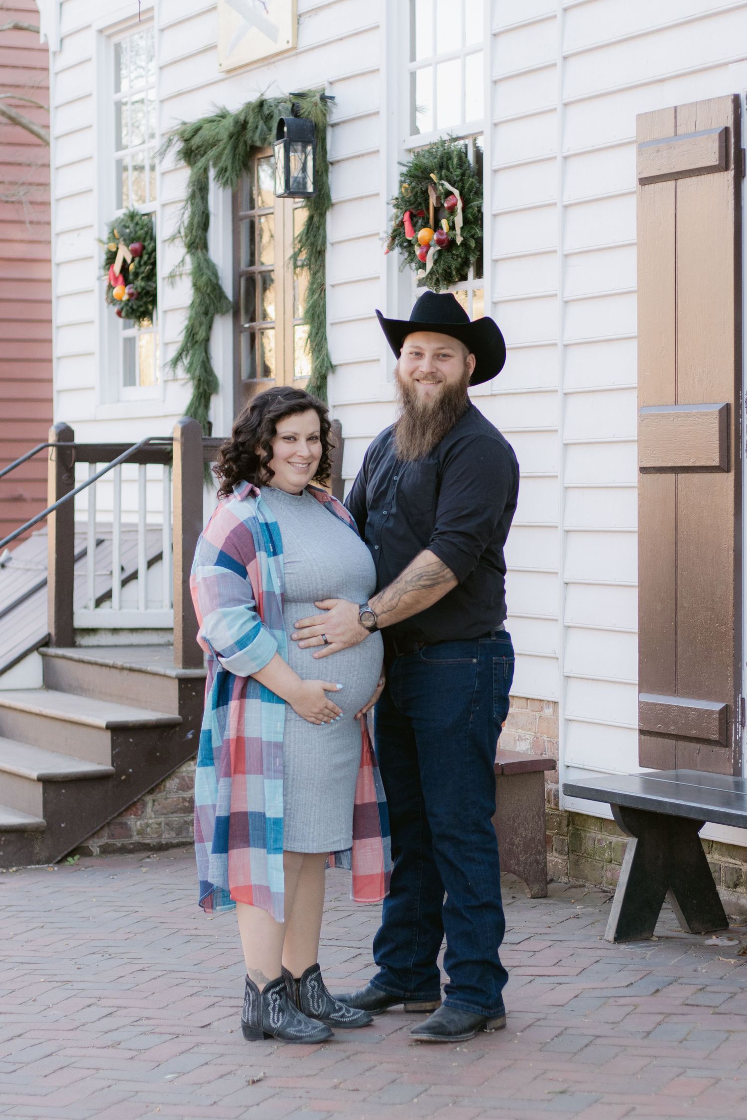 A couple in western attire embraces in front of a white building decorated with holiday garland and wreaths.