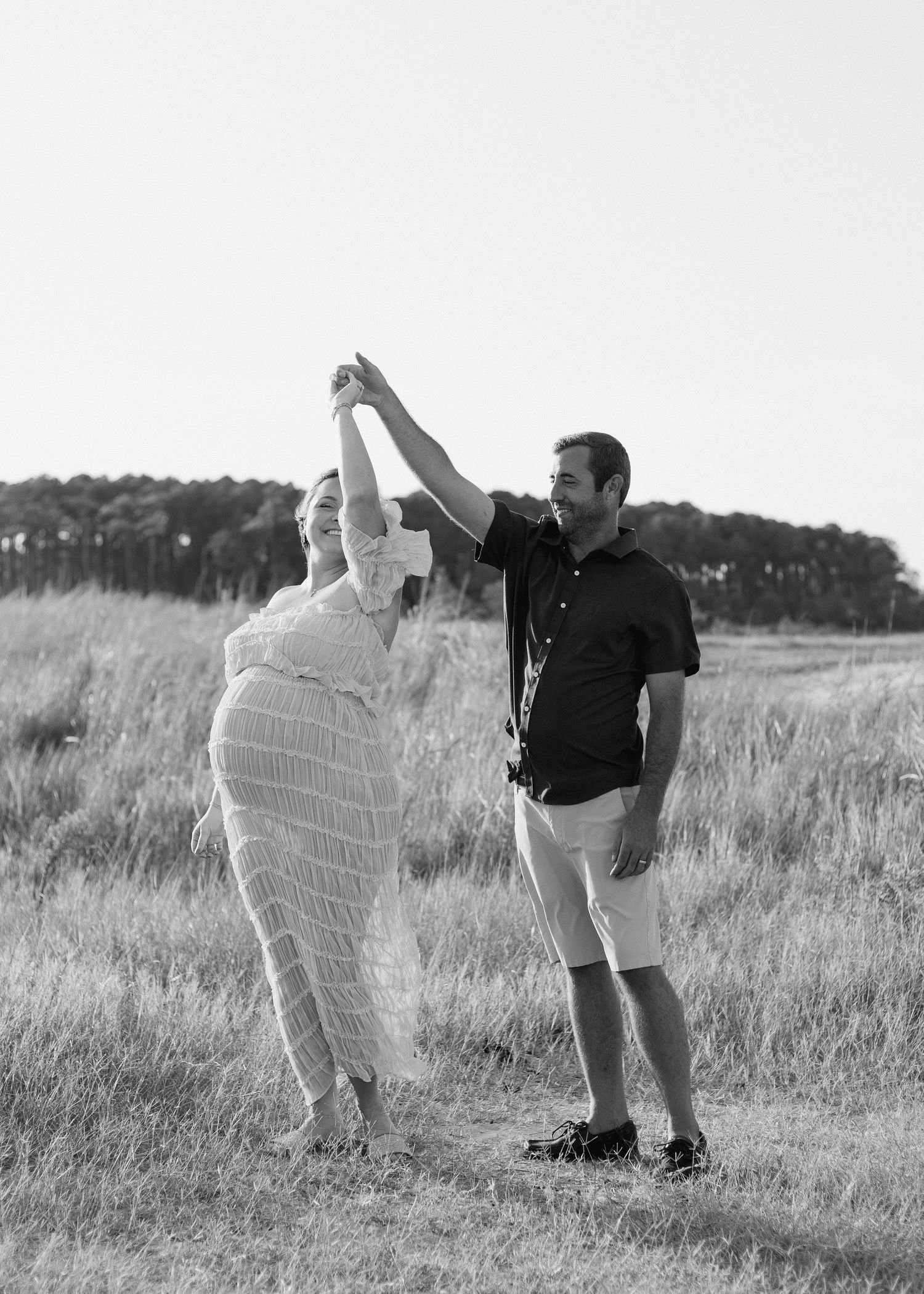 Black and white photo of a couple dancing outdoors in a field, man spinning woman in a white dress.