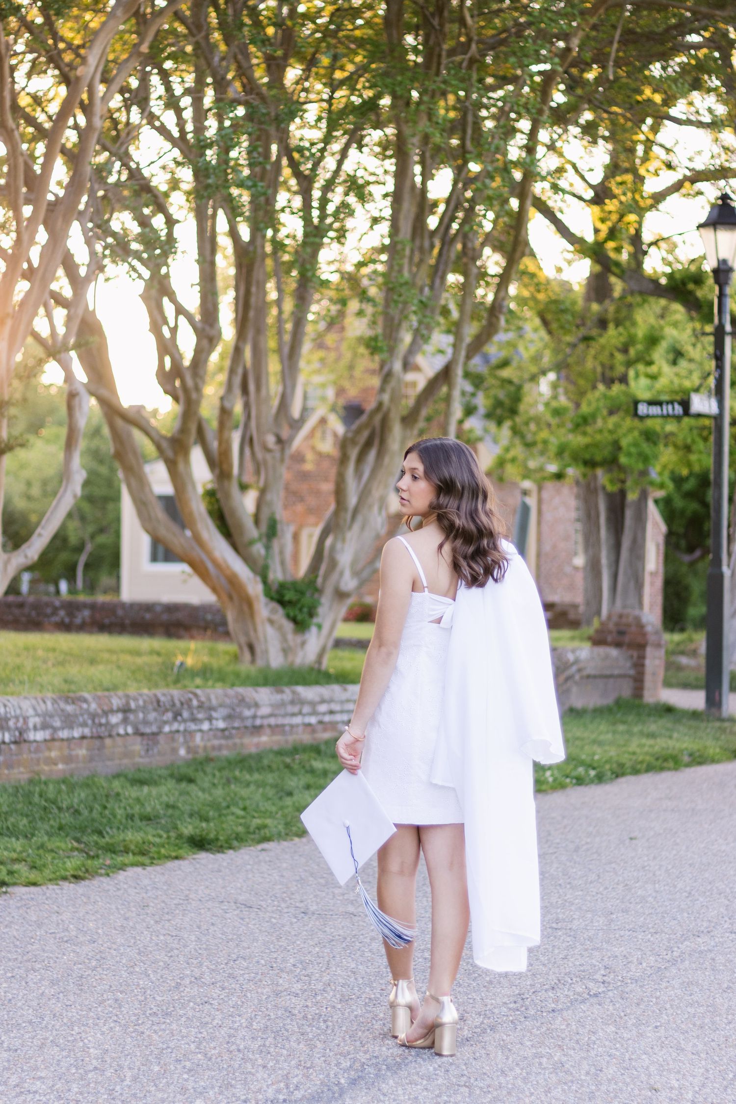 Two women walking down a tree-lined pathway wearing white dresses and sandals in a summer setting.