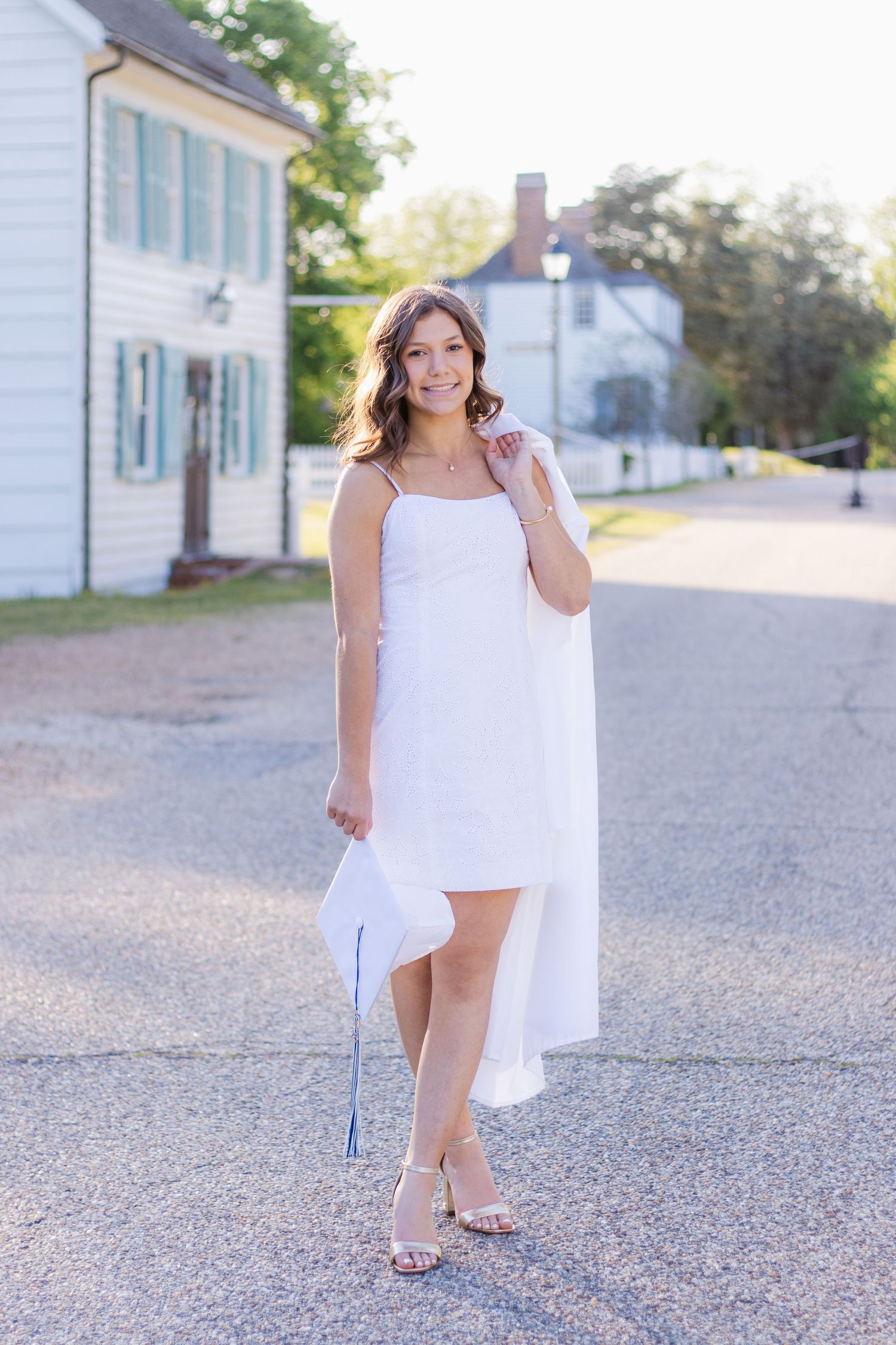 A woman in a flowing white dress poses gracefully on a gravel path near white colonial-style buildings on a sunny day.
