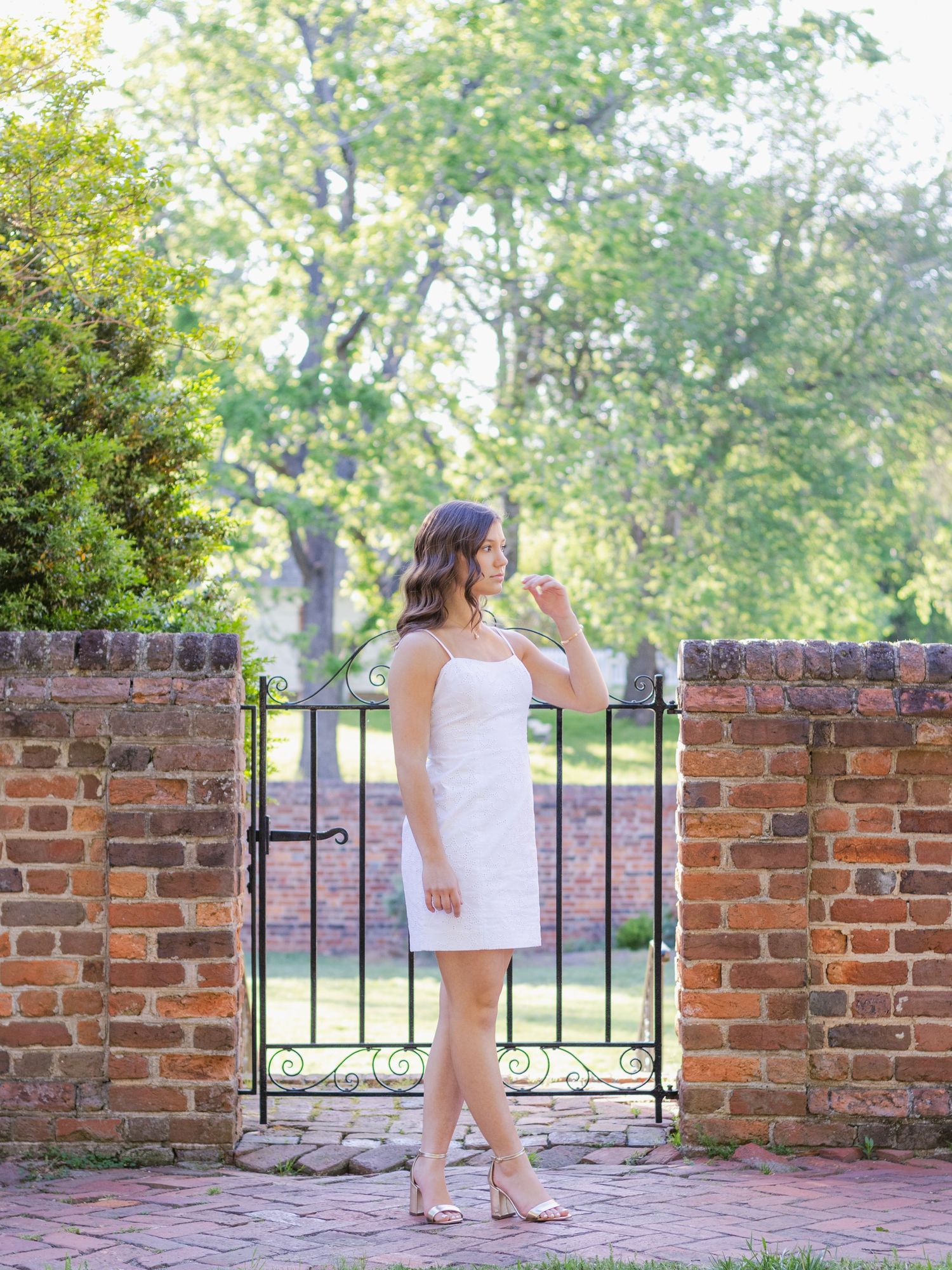 Woman in white strapless dress stands in front of rustic brick wall and iron gate on brick pathway in garden setting.