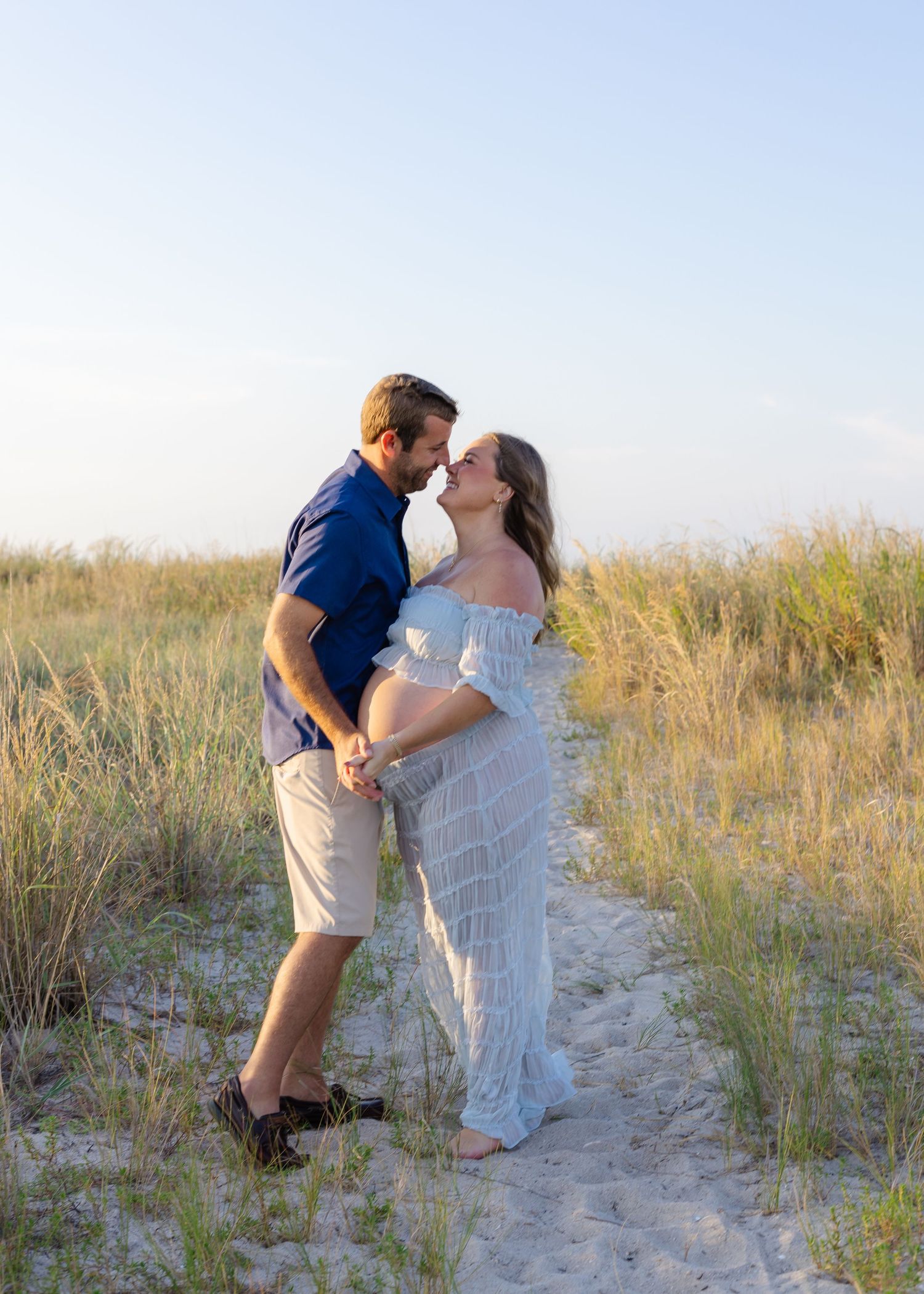 Couple sharing a tender moment on a sandy beach path, woman in white maternity dress, man in blue shirt, golden hour light.