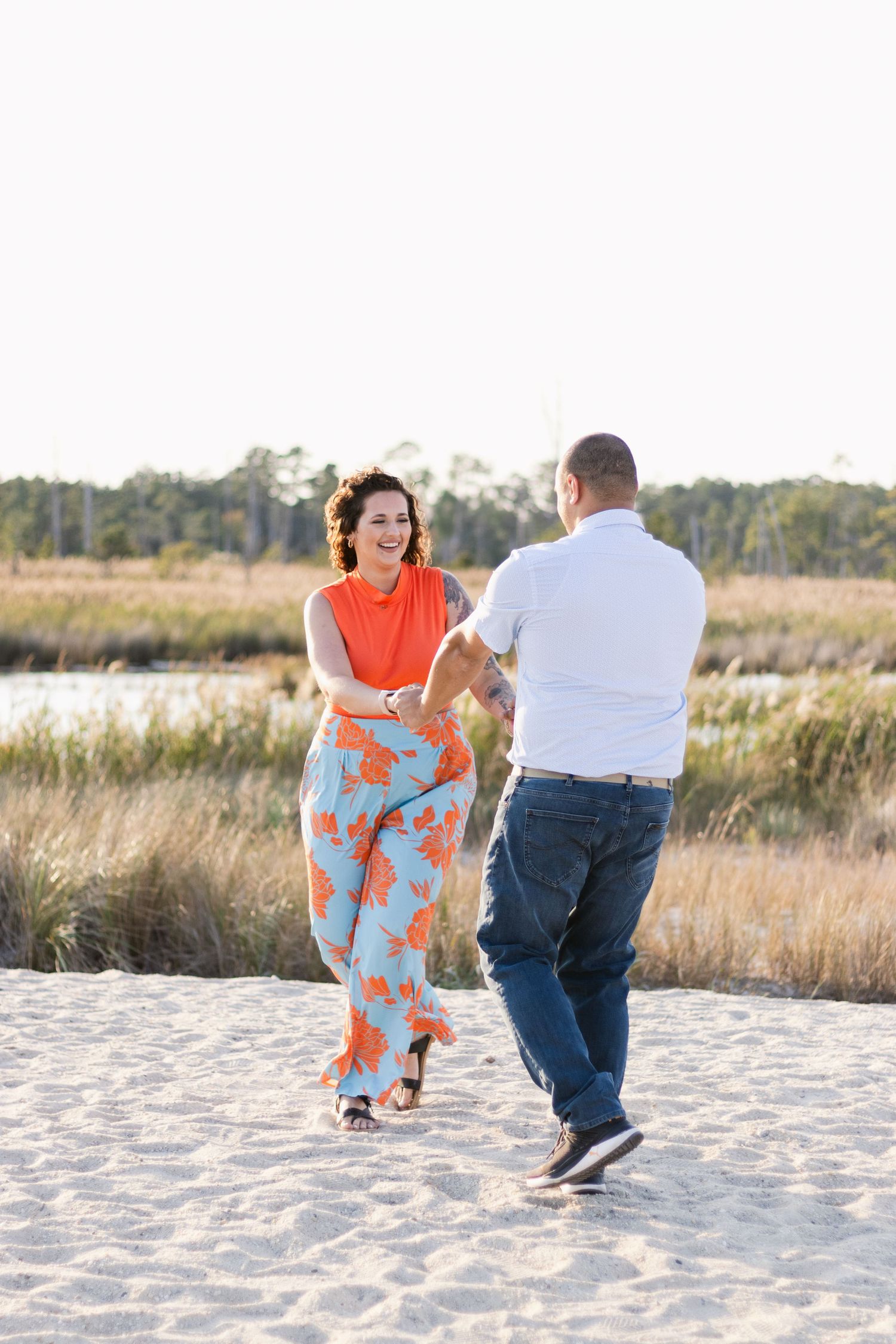 A couple dances playfully together on a white sandy beach with tall grass and trees in the background.