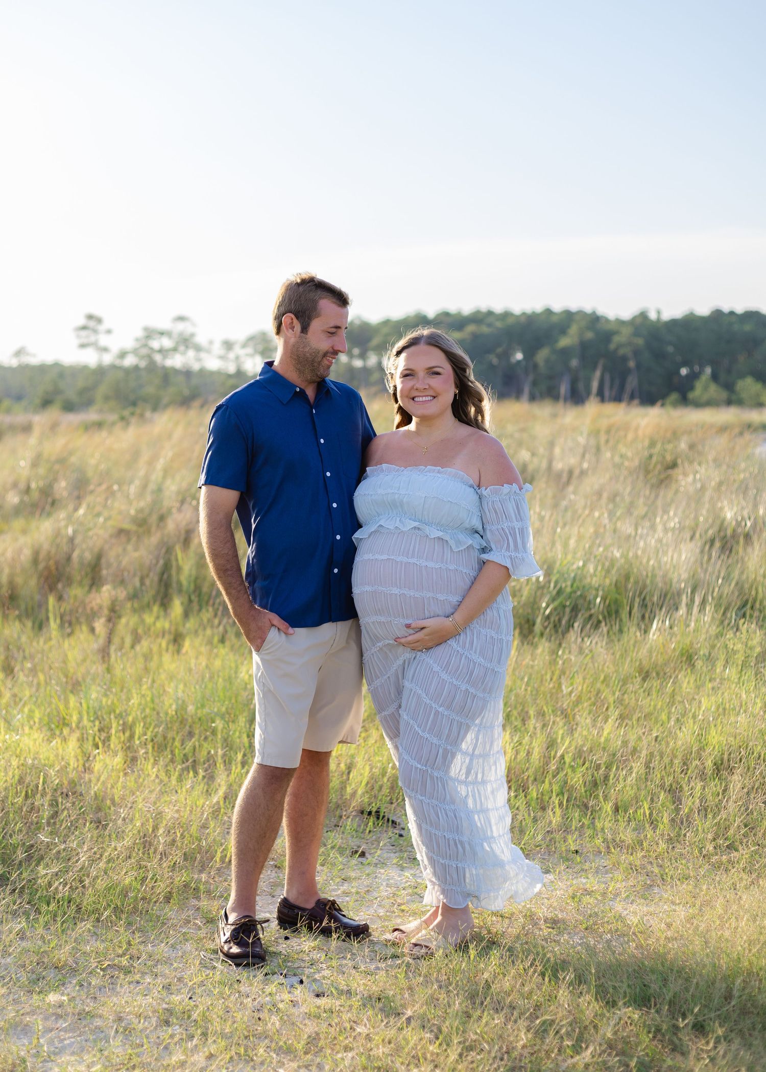 Smiling pregnant woman in white off-shoulder dress poses with man in blue shirt in sunny golden field.