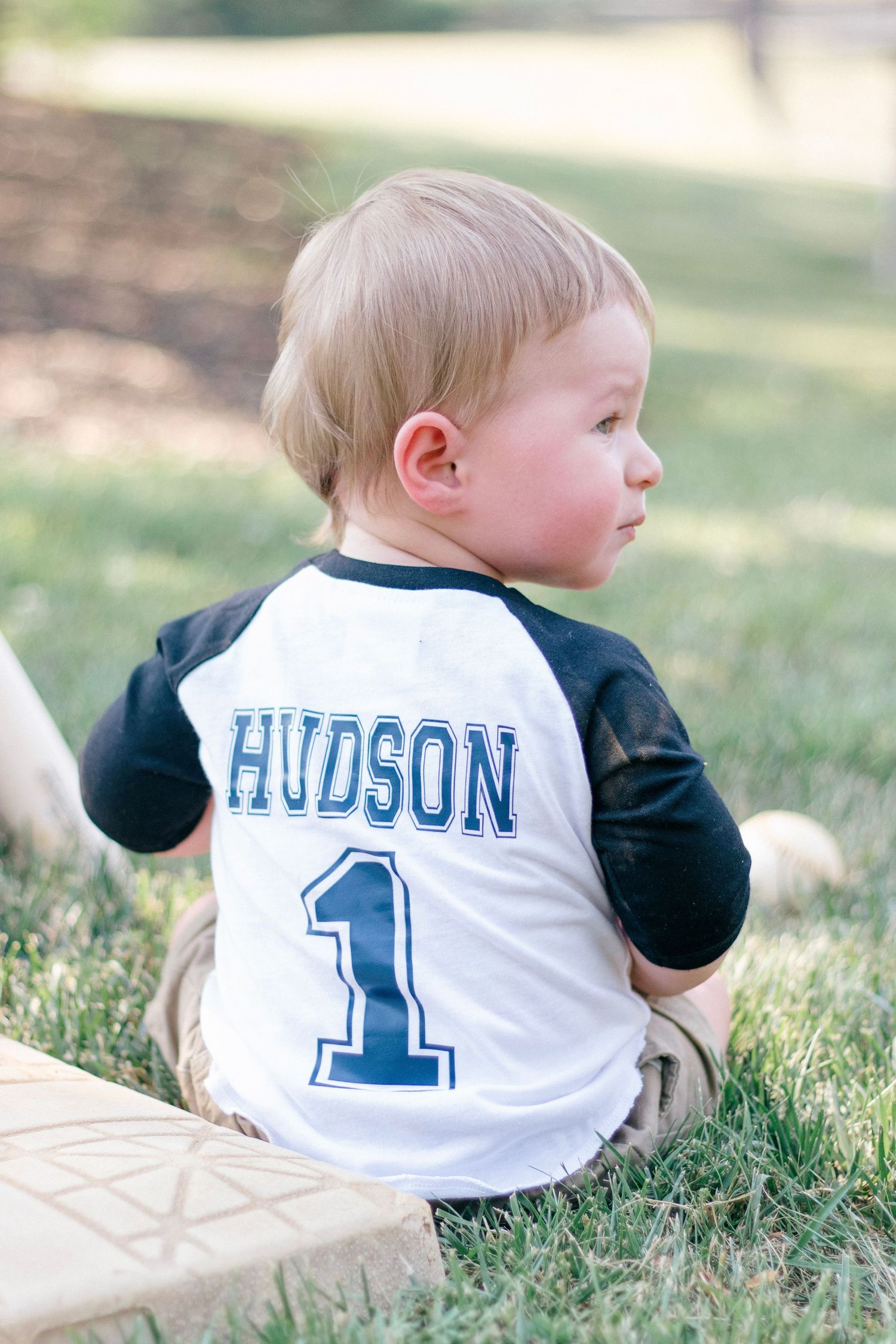 Baby wearing baseball style jersey with Hudson and number 1 printed on back lying on grass.