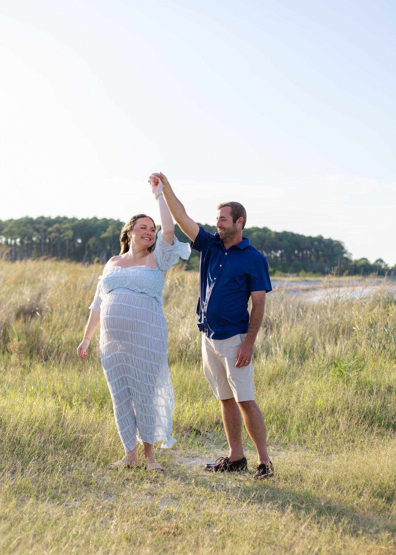 Pregnant woman in white dress dances with man in blue shirt in sunny golden field near water.
