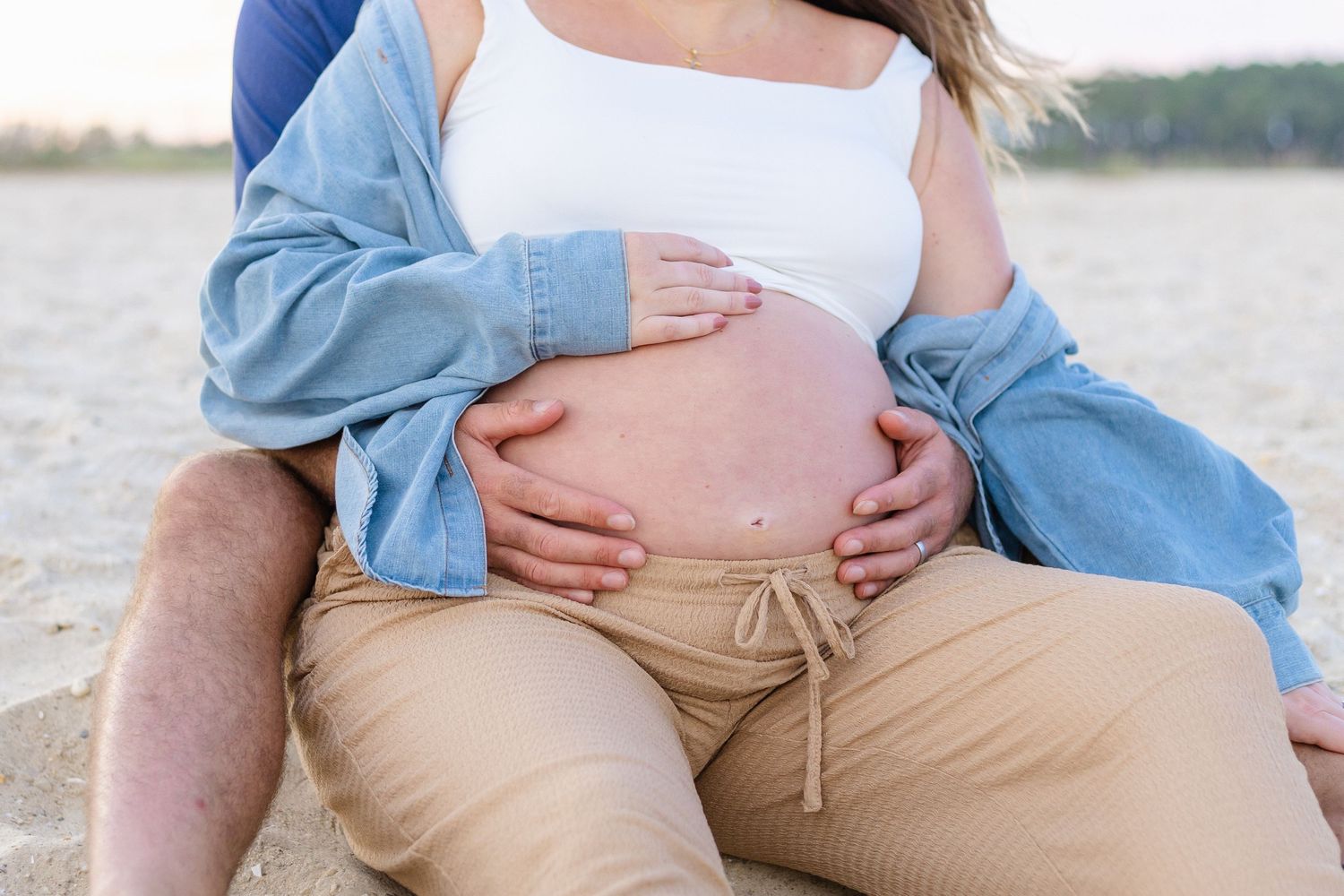 Pregnant woman sitting on beach with partner's hands cradling her bare baby bump outdoors.