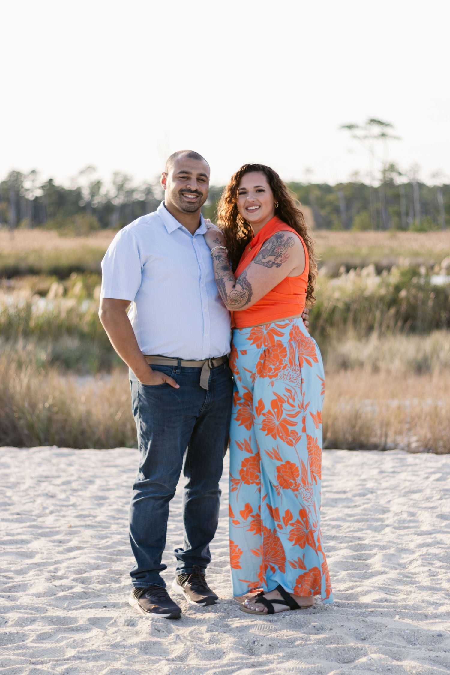 A couple poses together on a sandy beach at sunset with tall grass in the background.