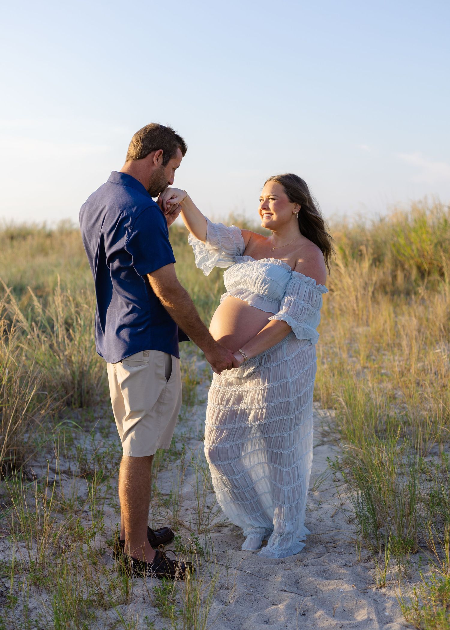 Couple dancing on beach sand dunes, pregnant woman in white lace dress holding hands with man in blue shirt.