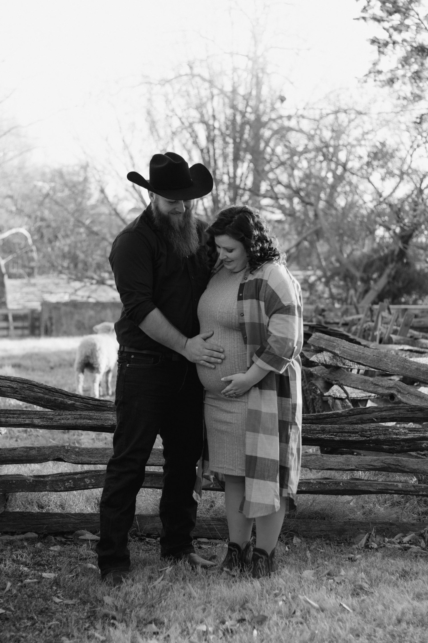Black and white portrait of an expectant couple in western attire standing by a rustic wooden fence in a countryside setting.