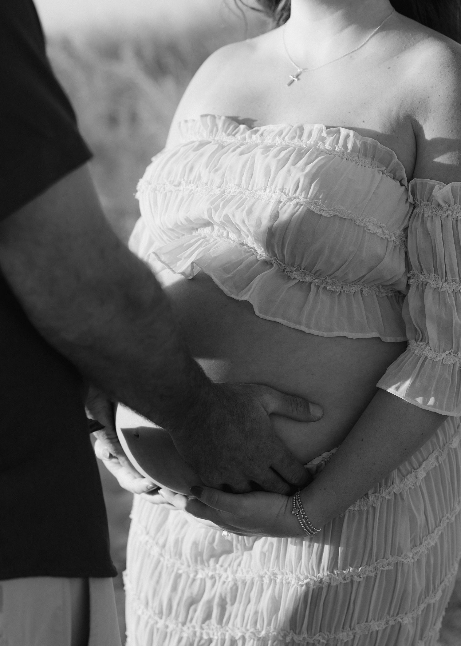 Black and white maternity photo of couple cradling pregnant belly, woman in ruffled off-shoulder white dress wearing cross necklace.