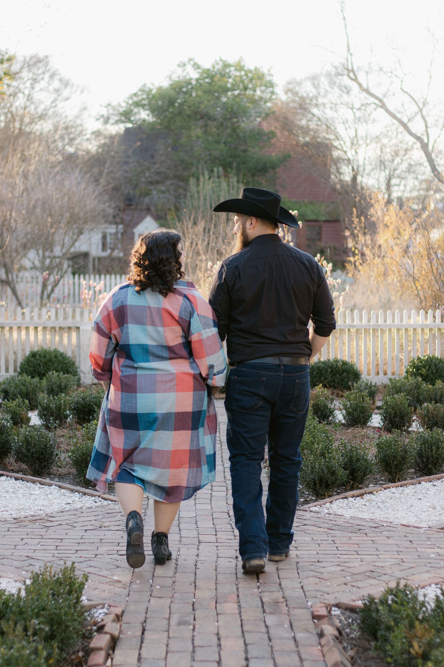 Two people in contrasting outfits walk together on a brick pathway through a garden setting at dusk.