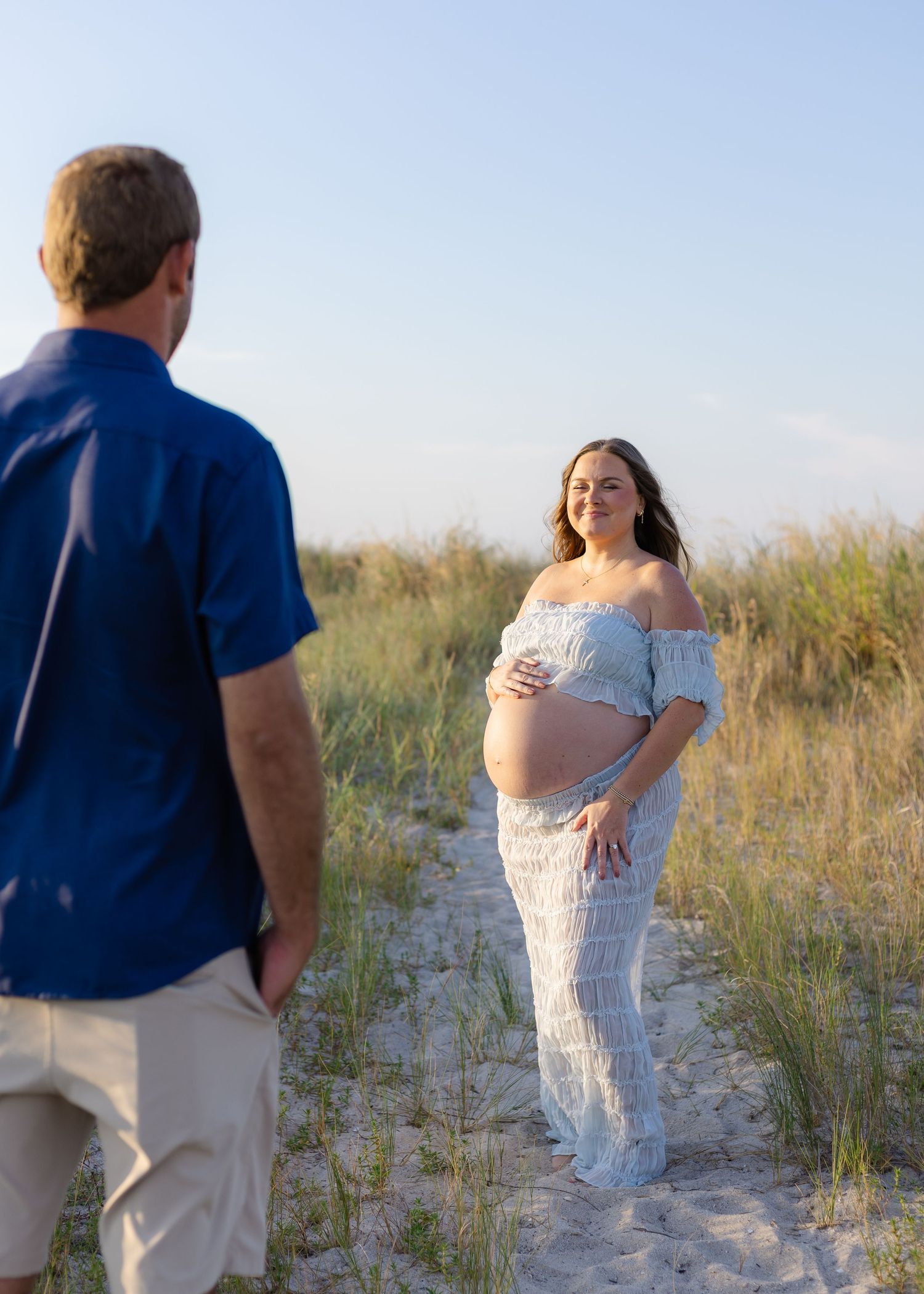 Pregnant woman in white lace dress holds baby bump on sandy beach path while partner looks on during maternity photo session.