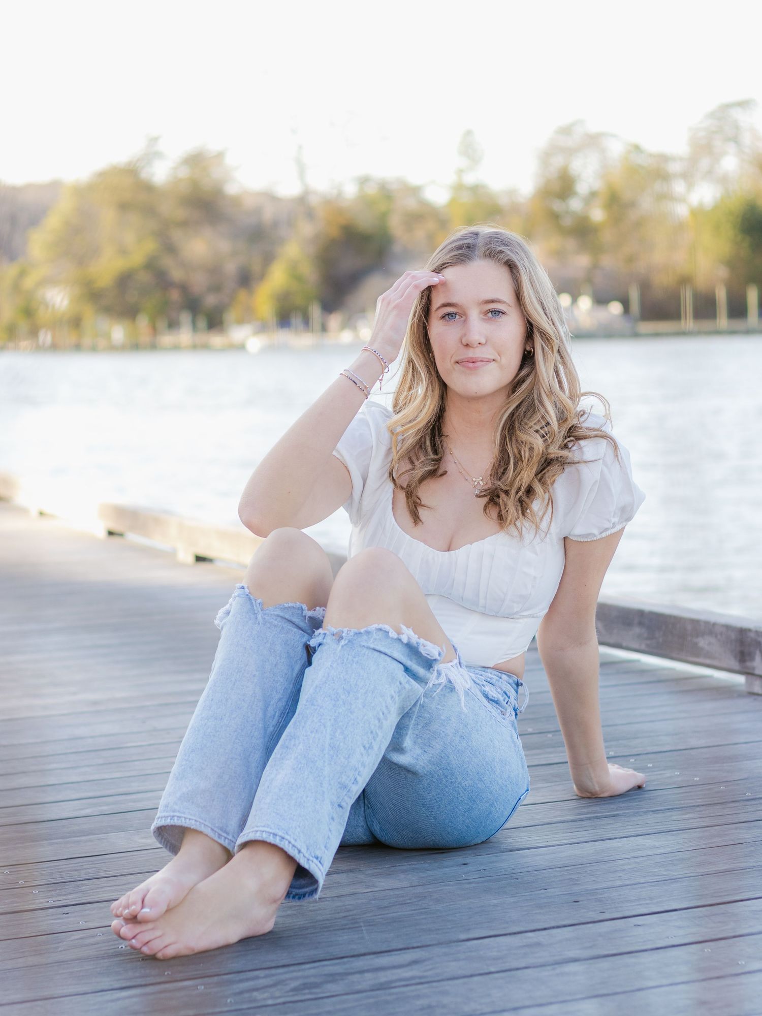 Young person in white top and jeans sits barefoot on wooden dock by serene lake on sunny autumn day.