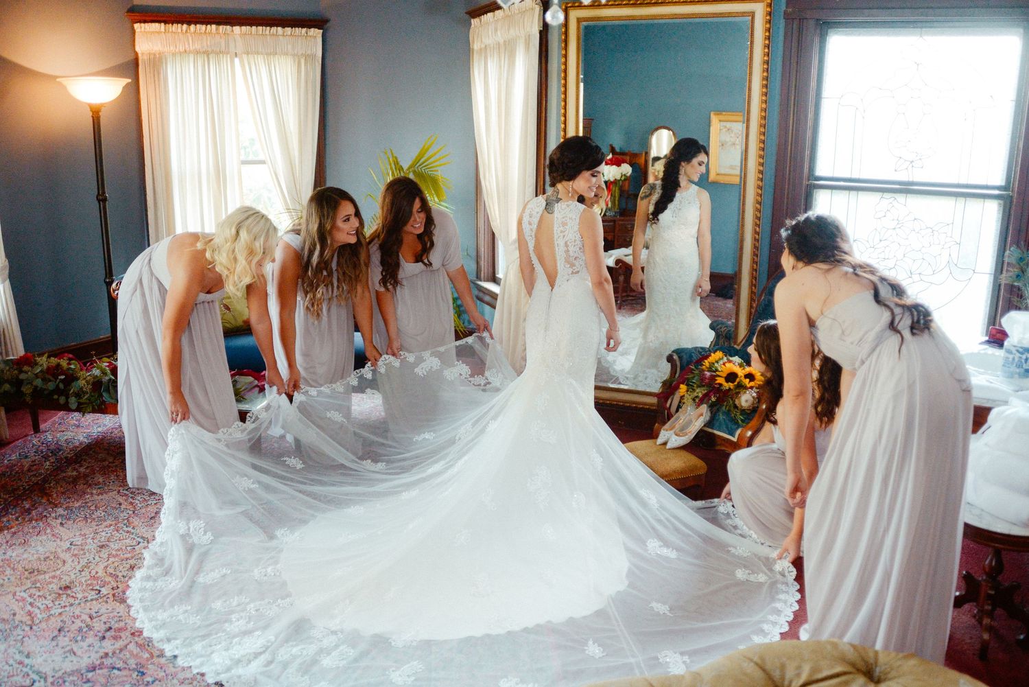 Bridesmaids in white dresses help arrange a wedding gown's long train on a patterned carpet.