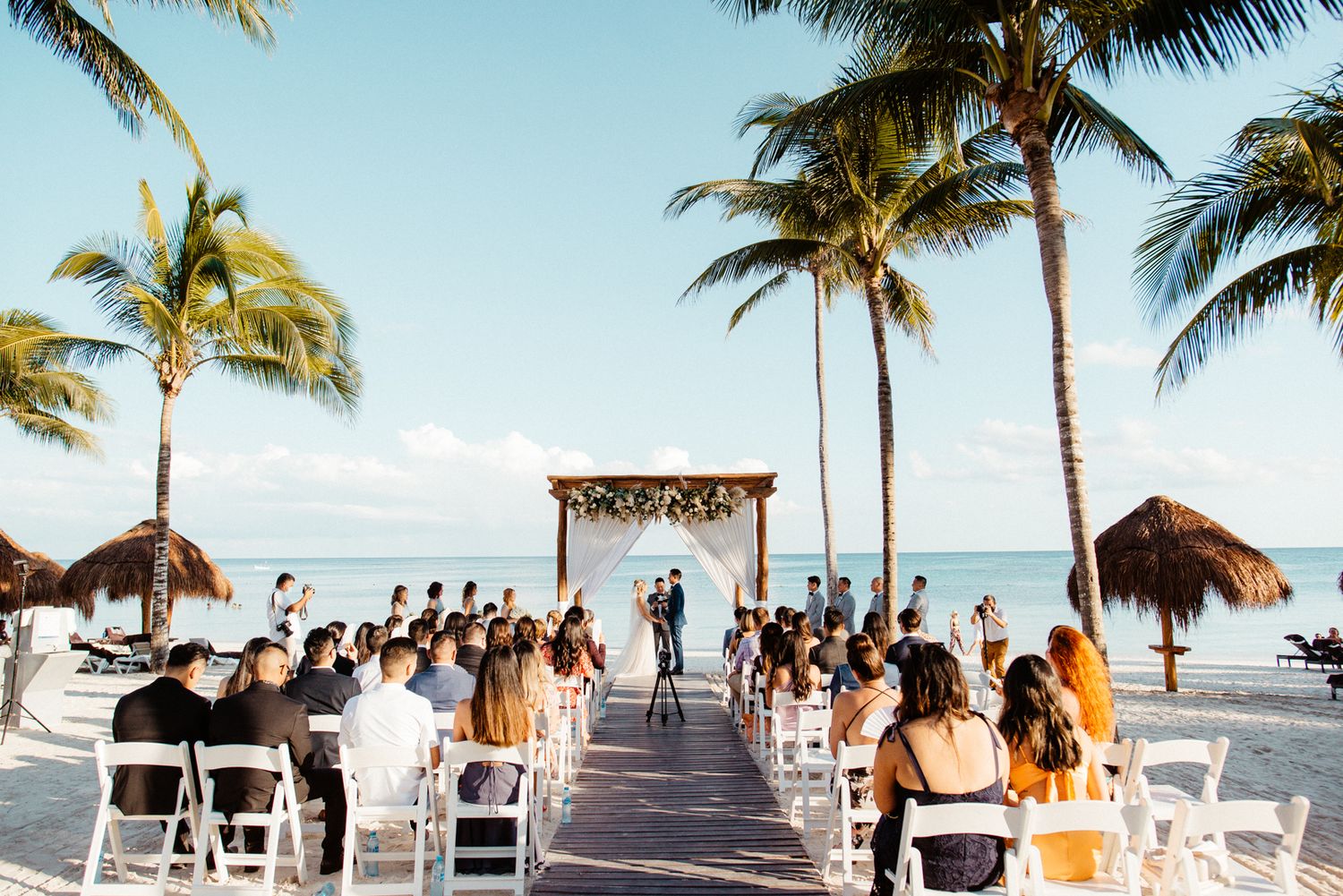 Outdoor beach wedding ceremony with guests seated in white chairs under palm trees near the ocean in Tulum, Mexico.