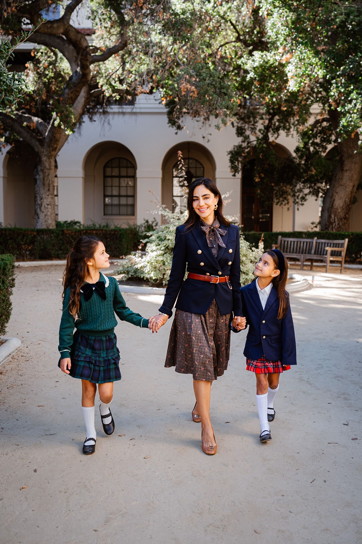 A woman in a navy blazer walks hand-in-hand with two girls in school uniforms through a sunlit courtyard.